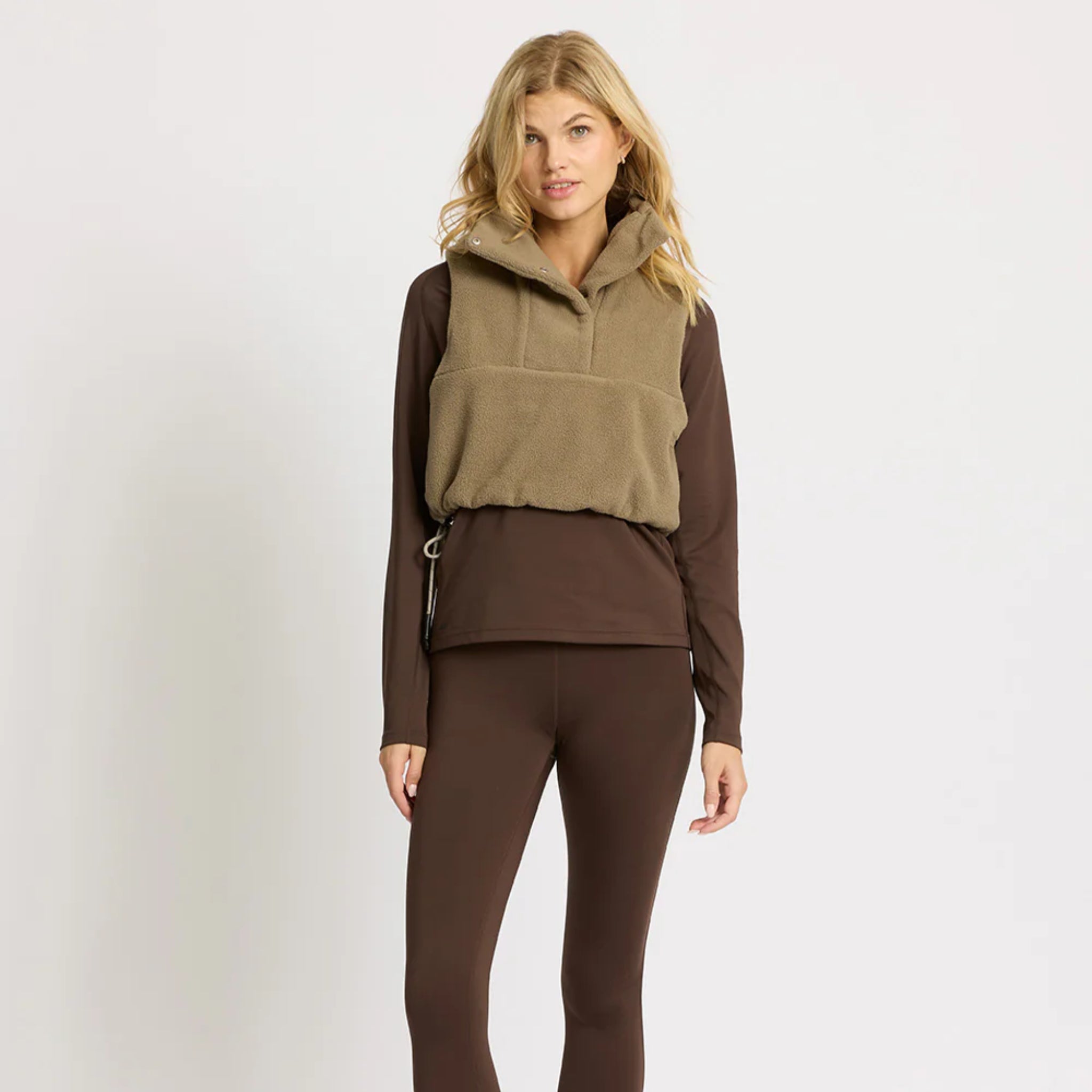 Eine Frau mit blondem Haar, die eine hellbraune Fleece-Weste über einem langärmeligen dunkelbraunen Oberteil und passenden Leggings trägt, steht vor einem schlichten hellen Hintergrund.