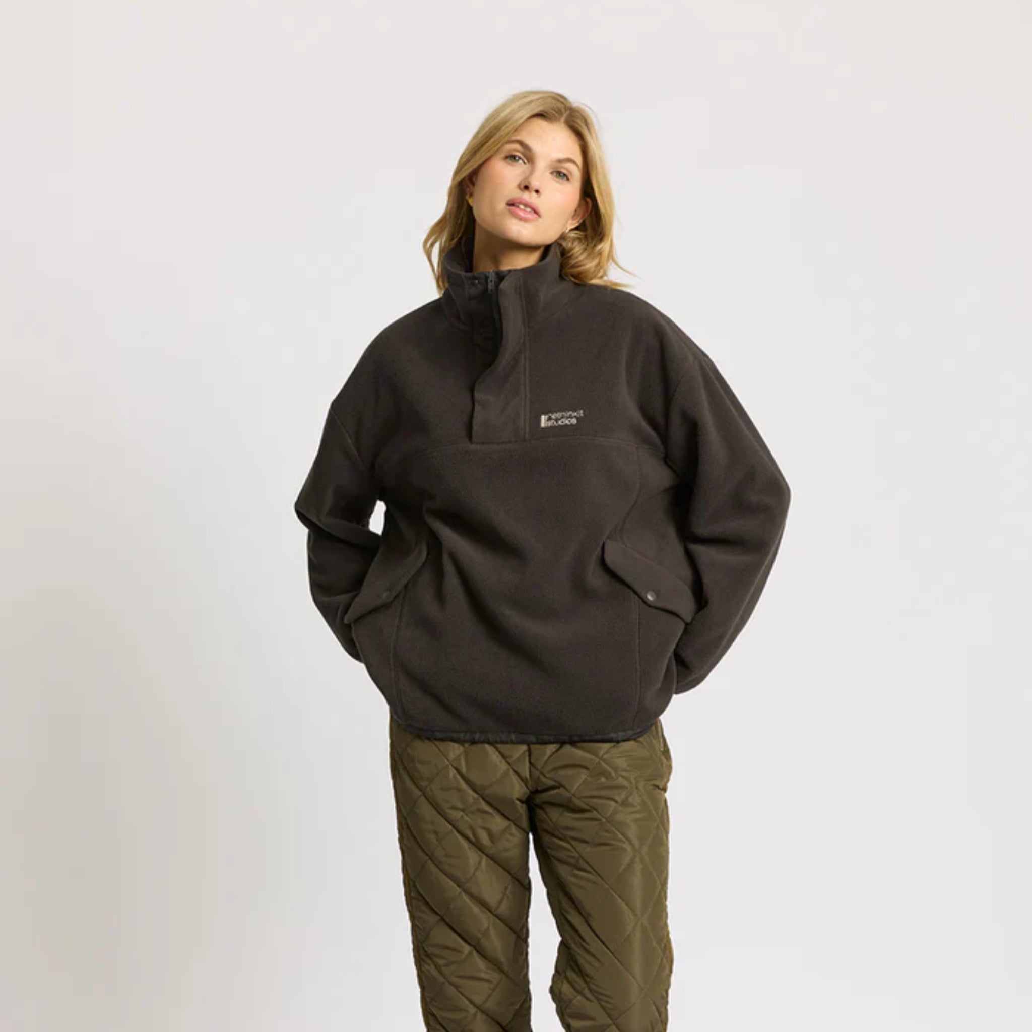 Eine Frau, die einen dunklen Fleecepullover und eine olivgrüne Stepphose trägt, steht vor einem schlichten hellen Hintergrund, hat die Hände in den Taschen und schaut leicht zur Seite.