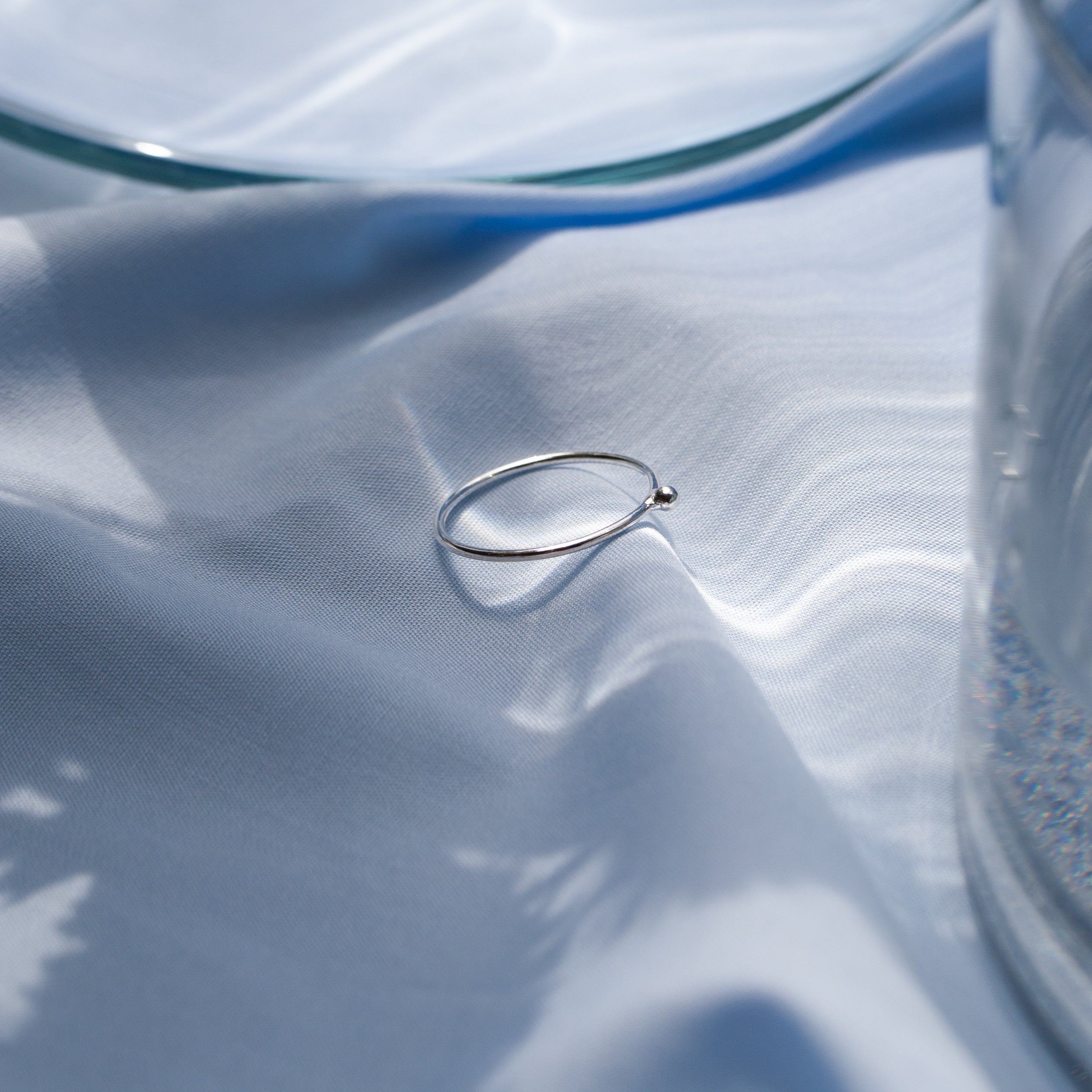 Ein silberner Ring liegt auf einem blauen Stoff unter einer Glasplatte im Sonnenlicht.