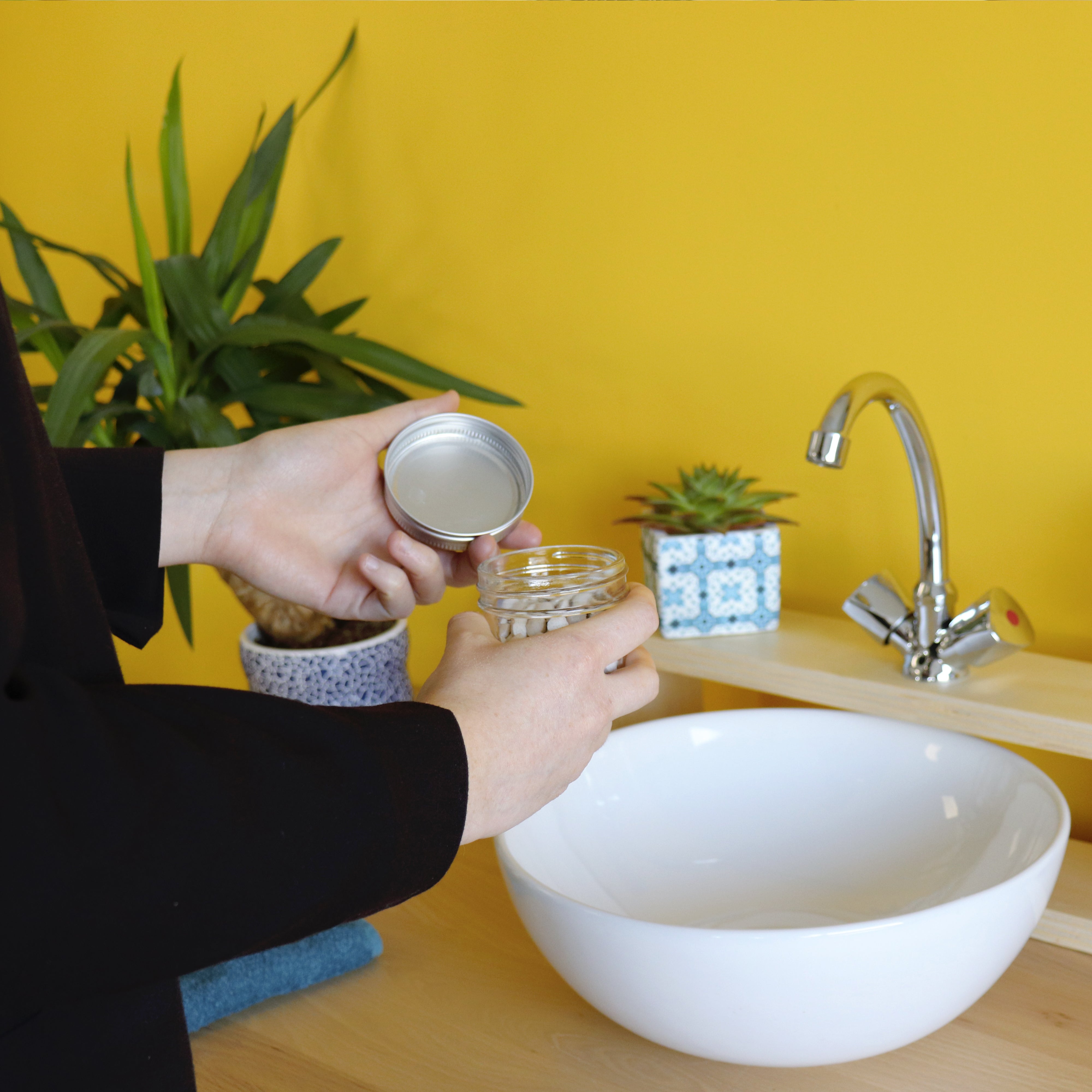 Eine Person öffnet ein Glasbehälter neben einem Waschbecken, mit Pflanzen im Hintergrund vor einer gelben Wand.