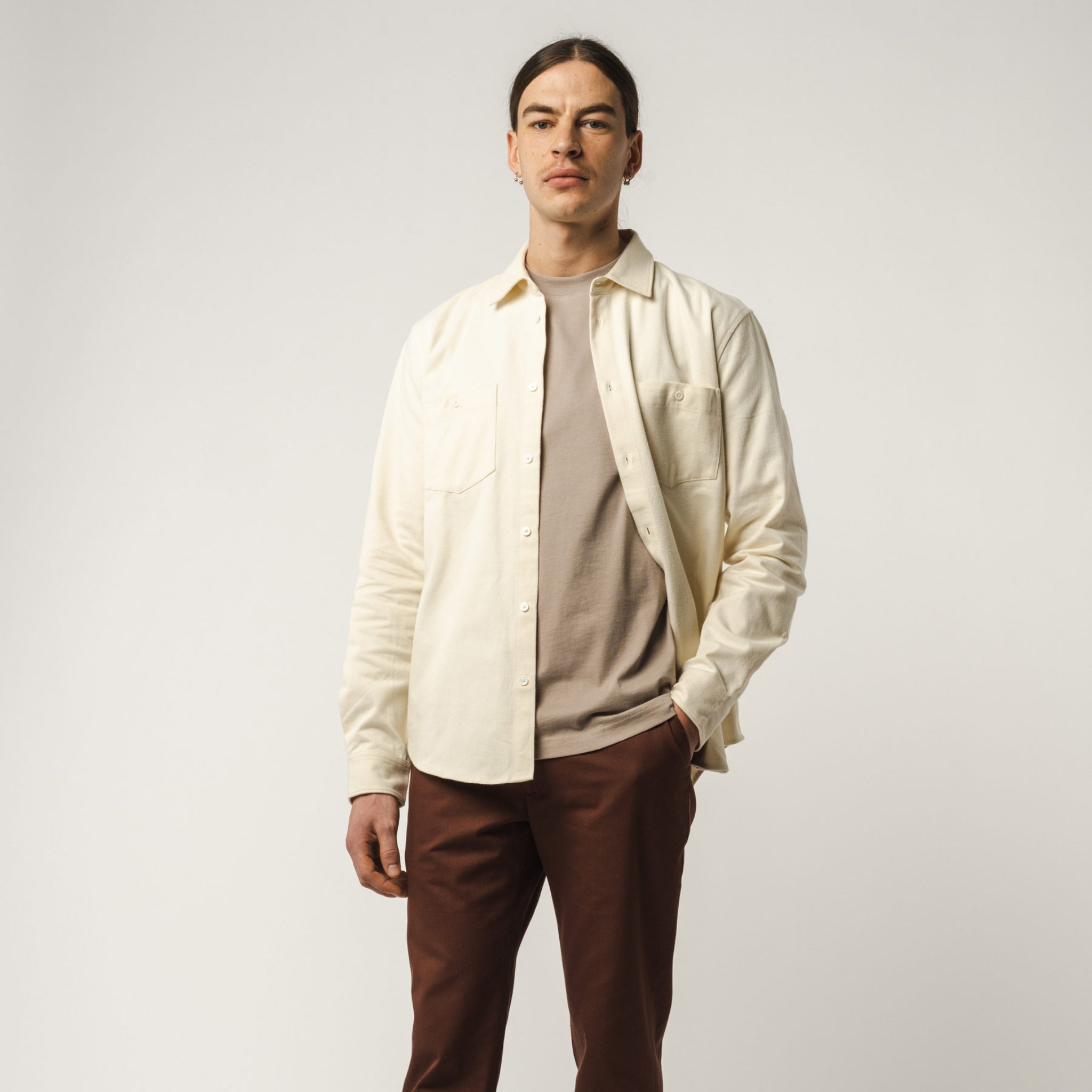 Mann in beiger Jacke und braunem Hose steht vor neutralem Hintergrund.