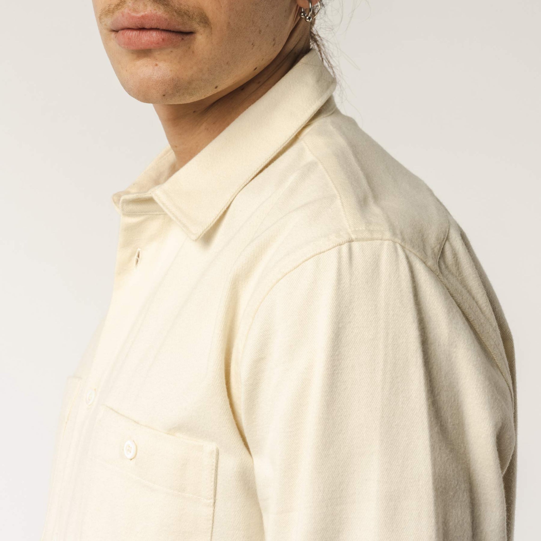 Eine Person in einem cremefarbenen Hemd, fokussiert auf die Schulterpartie und einen Teil des Gesichts mit Ohrringen.