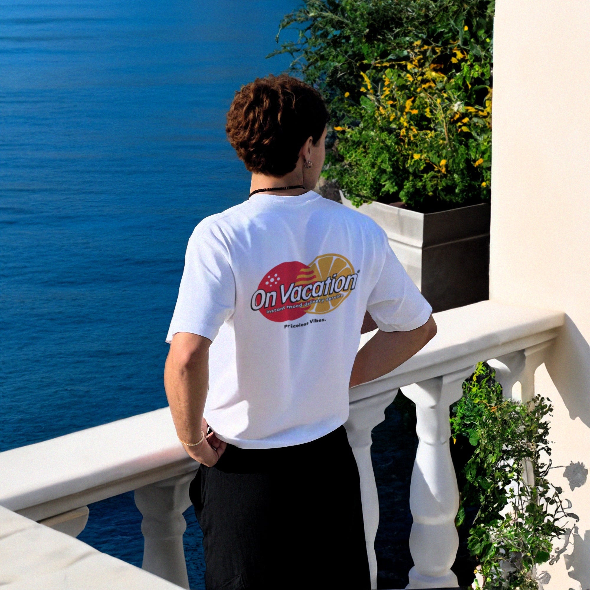 Eine Person mit kurzen Haaren, die ein weißes On Vacation-T-Shirt trägt, steht auf einem Balkon mit Blick auf ein blaues Meer mit Klippen und Grün im Hintergrund.