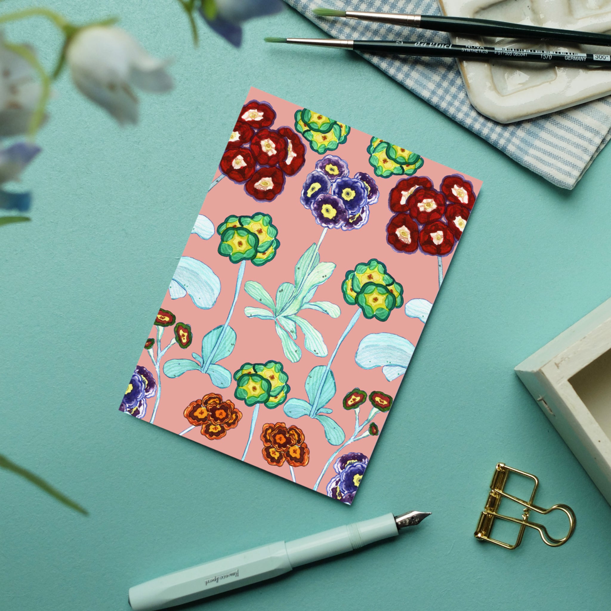 Illustration einer Blumenkarte auf blauem Tisch, umgeben von Pinsel, Notizbuch, Stift und goldener Büroklammer.