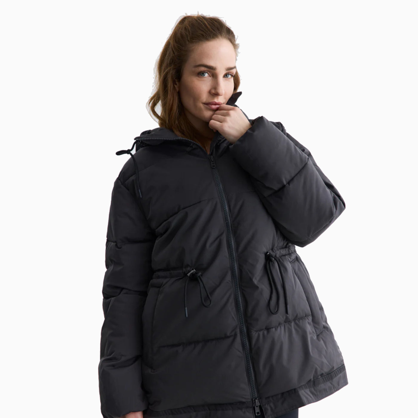 Eine Frau mit hellbraunem Haar, die eine schwarze Pufferjacke trägt, steht vor einem weißen Hintergrund, berührt mit einer Hand ihre Lippen und lächelt leicht.