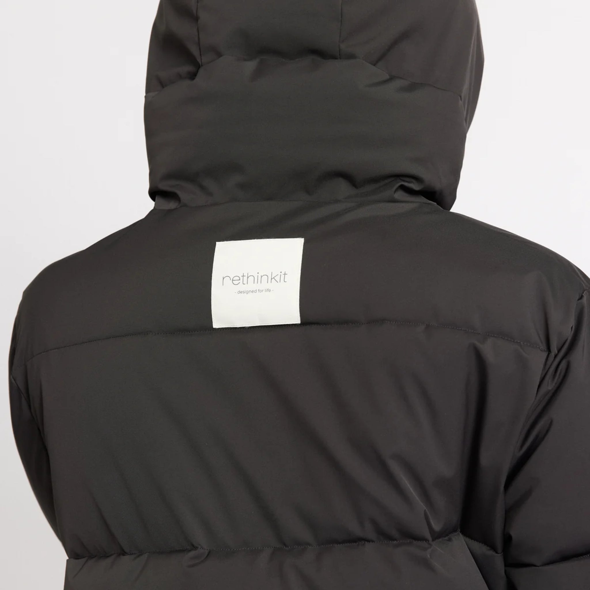 Eine Person, die eine bauschige schwarze Jacke mit einem weißen rechteckigen Etikett auf der Rückseite trägt, auf dem "rethinkit" steht. Die Jacke hat einen hohen Kragen und der Hintergrund ist einfarbig hellgrau.