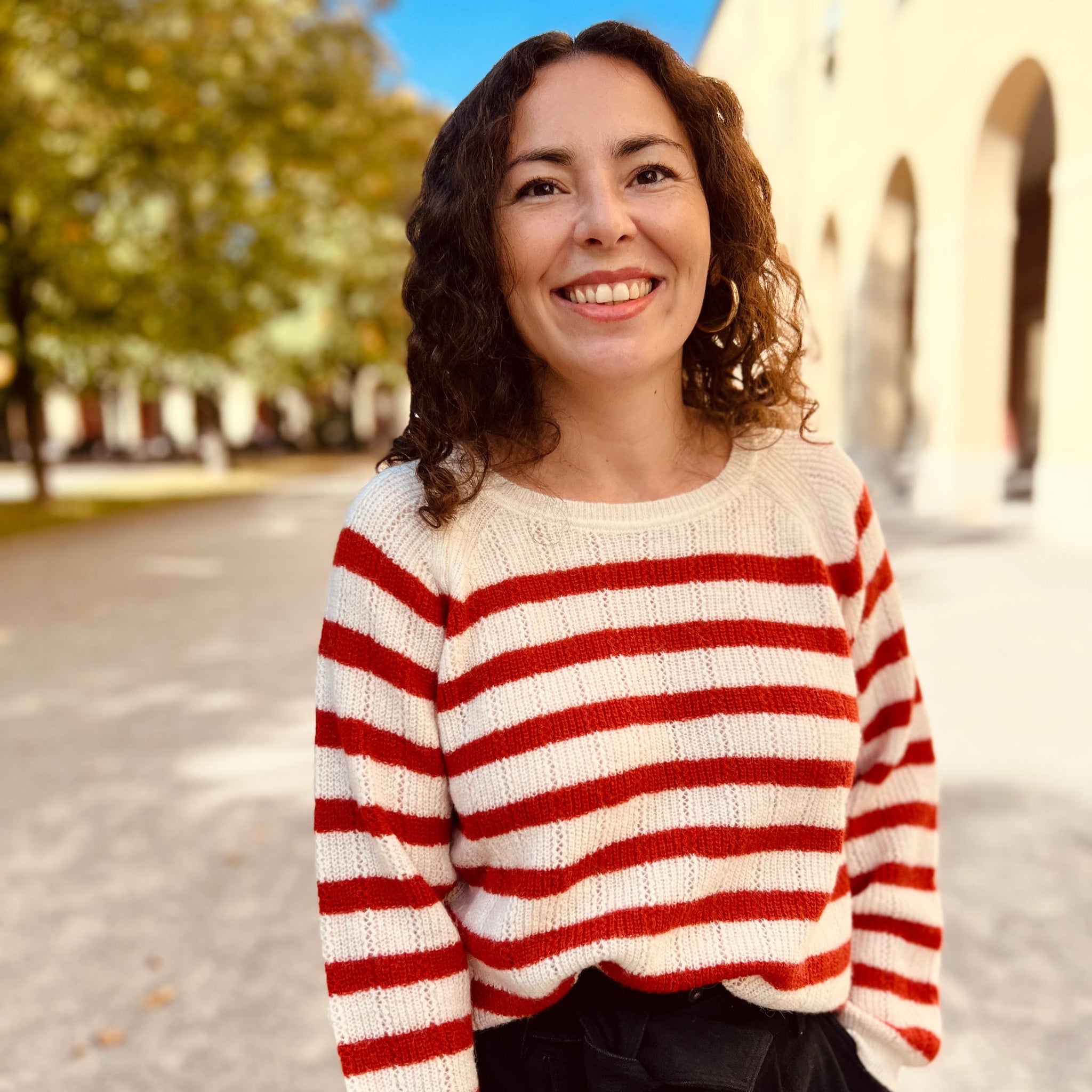 Eine Frau mit lockigem Haar trägt einen rot-weiß gestreiften Pullover und lächelt in einem sonnigen, parkähnlichen Umfeld.