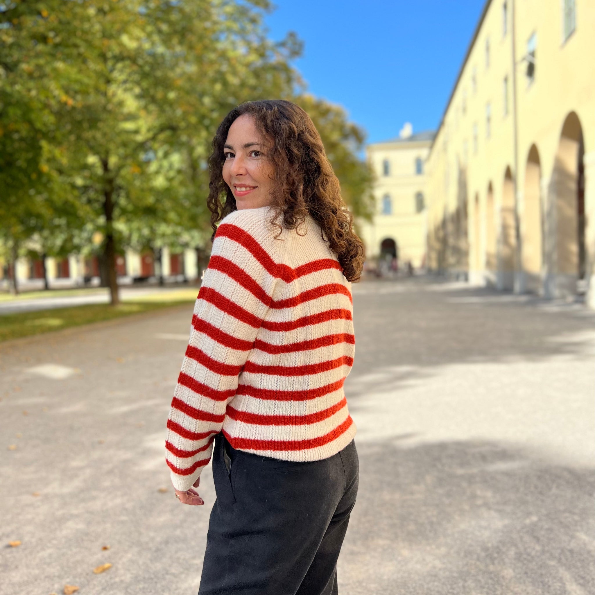 Frau mit lockigem Haar und gestreiftem Pullover steht draußen in der Sonne und lächelt.