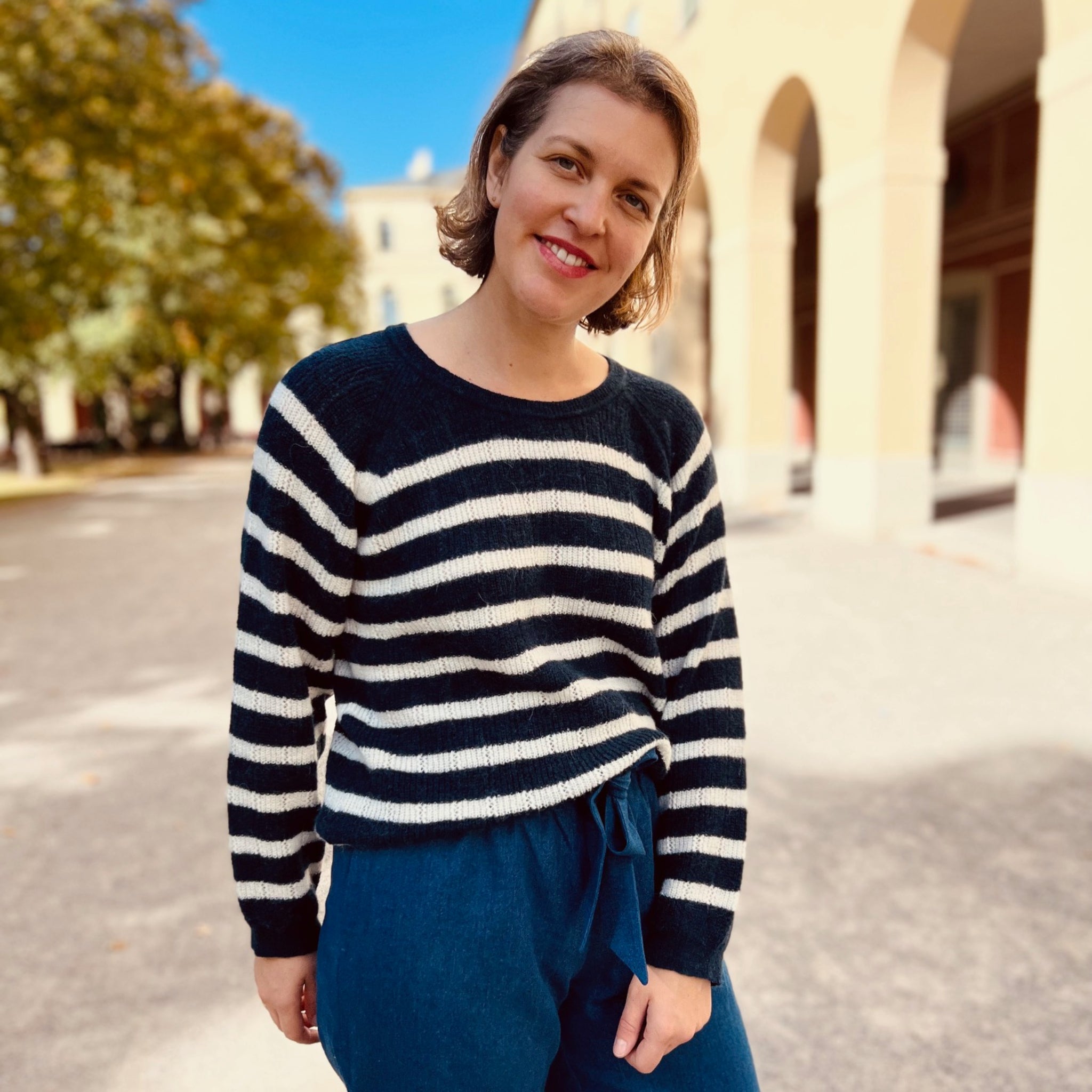 Frau in gestreiftem Pullover steht draußen vor einem Gebäude mit Bögen an einem sonnigen Tag.