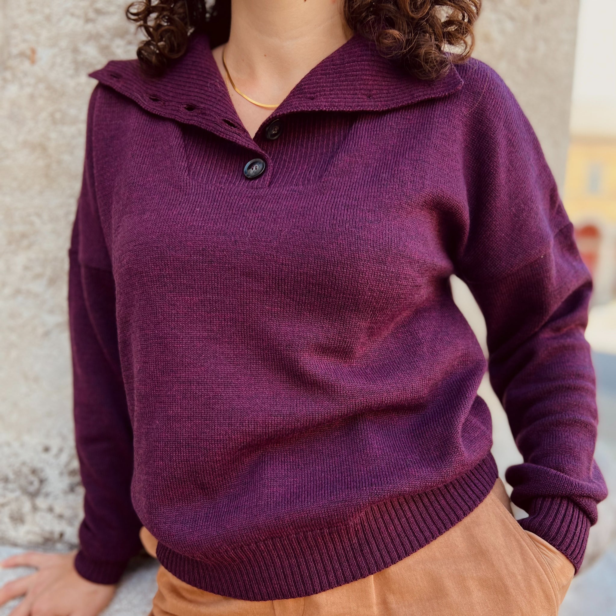 Person in violettem Pullover mit Polokragen und brauner Hose lehnt an einer Wand.