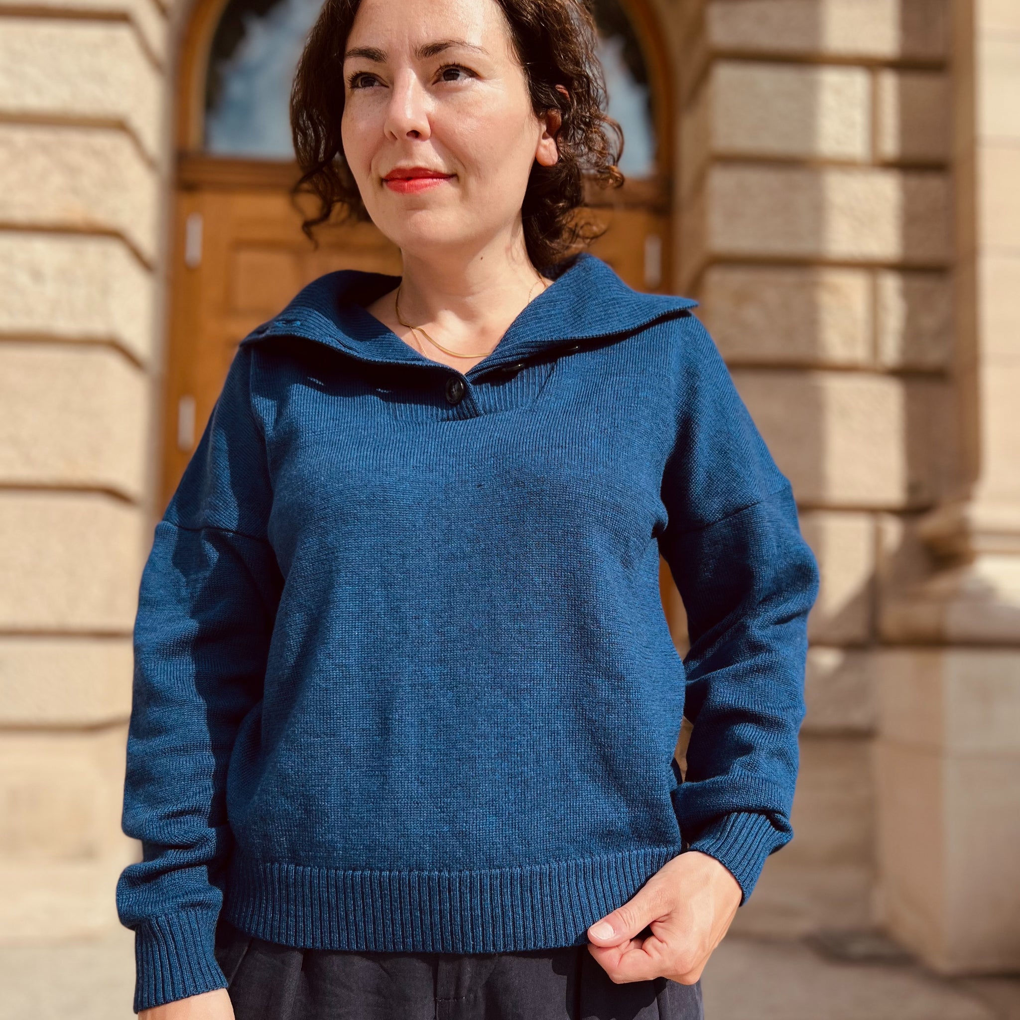Eine Frau trägt einen blauen Pullover und steht vor einem Gebäude mit steinernen Säulen und Holztüren.