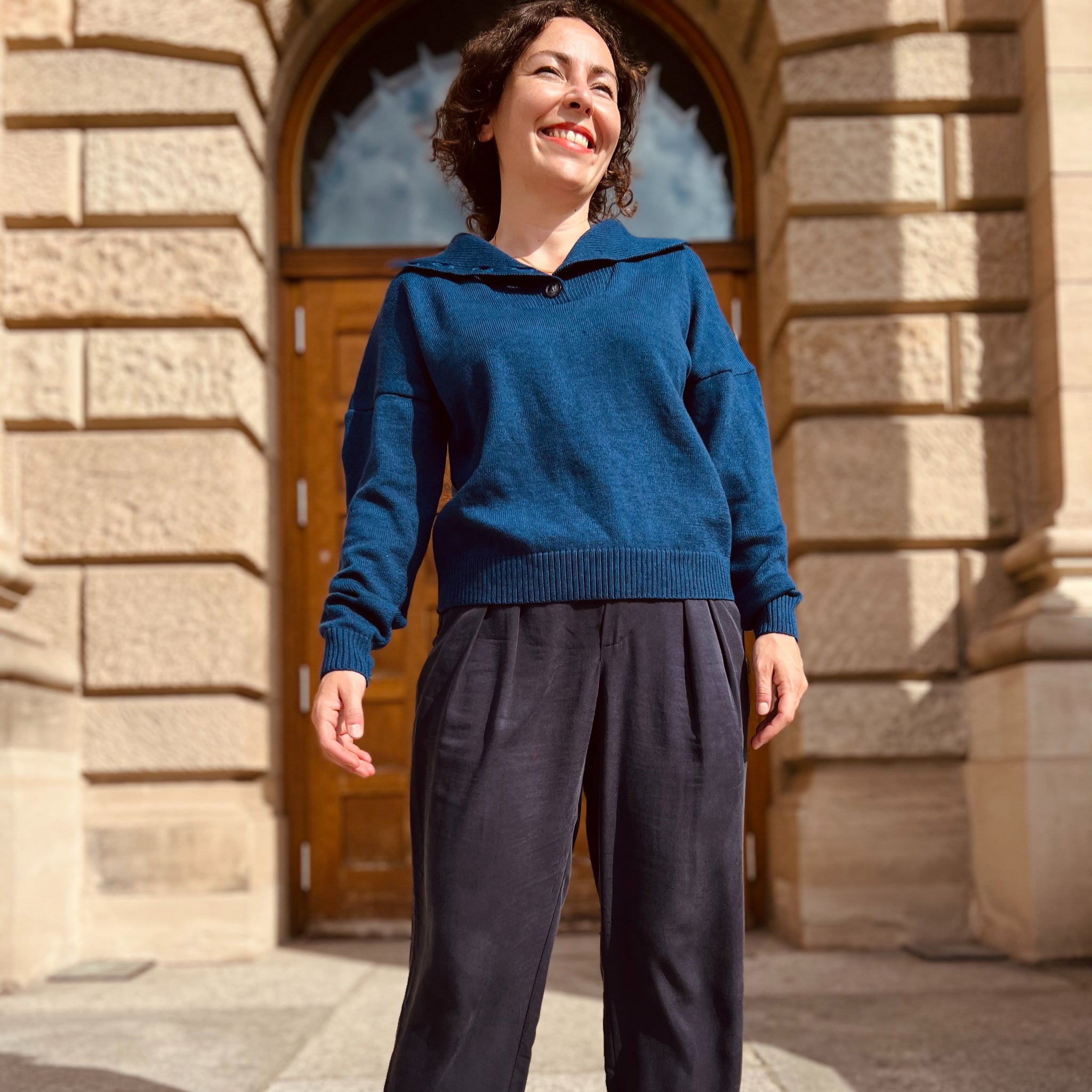 Frau in einem blauen Pullover und schwarzen Hosen steht lächelnd vor einem Gebäude mit Holztür und Steinwänden.