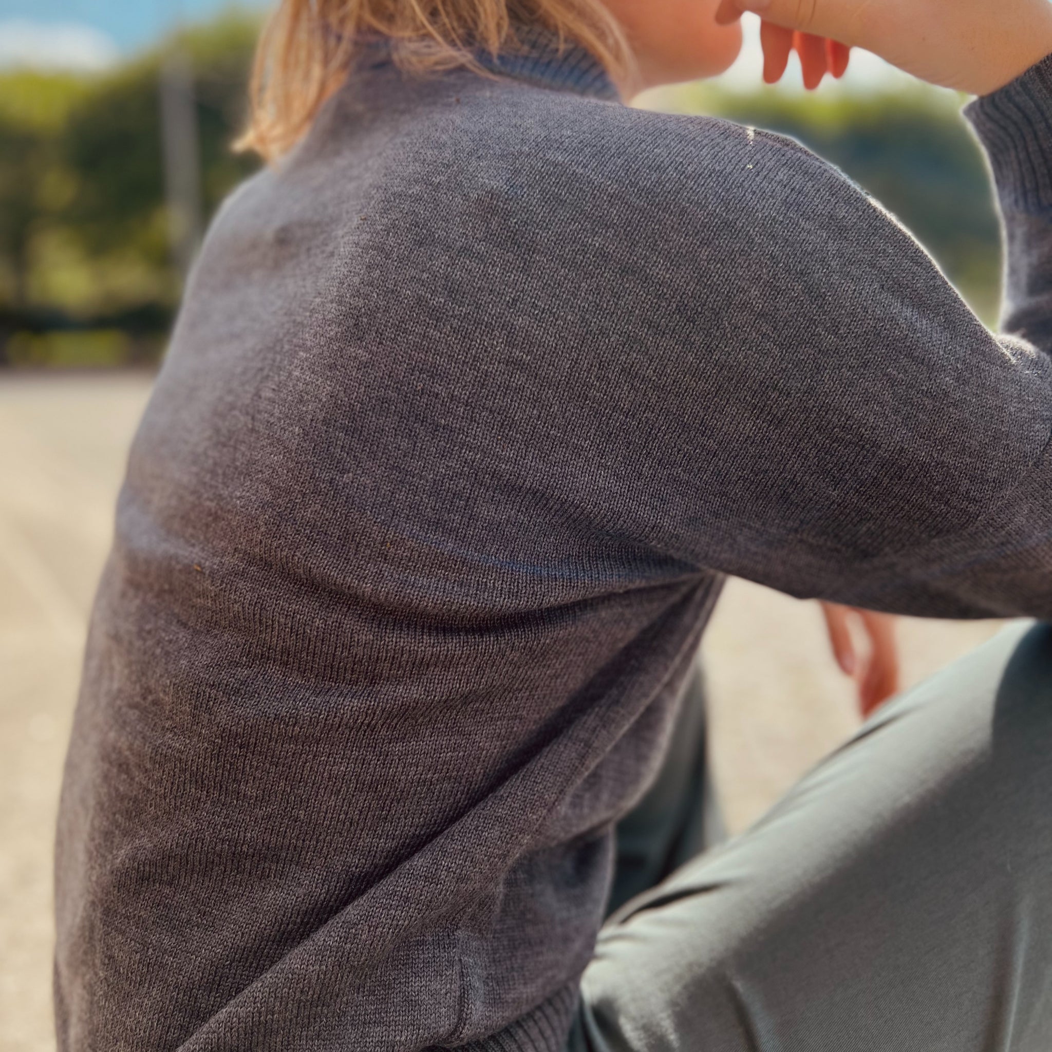 Person mit grauem Pullover sitzt draußen in der Sonne, die Hand am Kinn, unscharfer Hintergrund mit Bäumen.