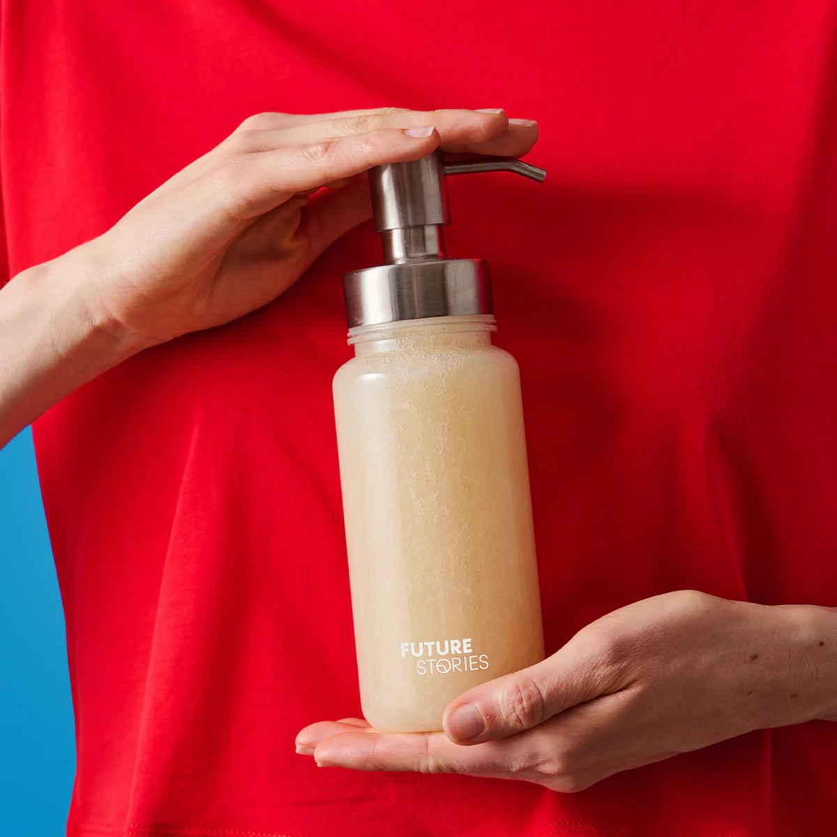 Eine Person in einem roten Hemd drückt auf die Pumpe einer durchsichtigen Flasche, die mit einer blassen Flüssigkeit gefüllt ist und die Aufschrift FUTURE STORIES trägt, und hält sie mit der anderen Hand sanft vor einem blauen Hintergrund.