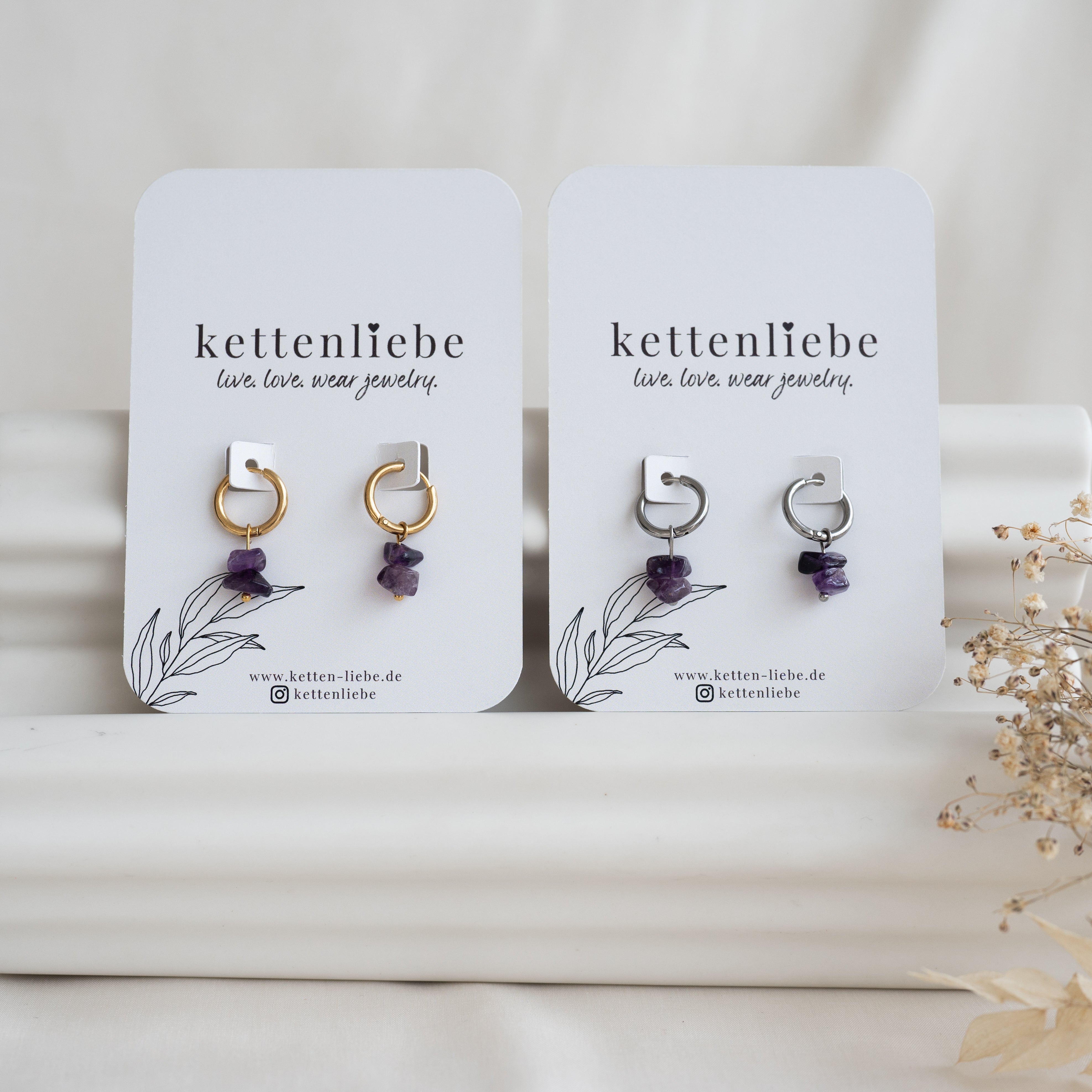 Zwei Paar Ohrringe auf weißen Karten mit der Aufschrift "kettenliebe". Jedes Paar besteht aus einem Reif - einer aus Gold, einer aus Silber - mit einem kleinen violetten Edelsteinanhänger, der vor einem weichen Hintergrund mit getrockneten Blumen steht.