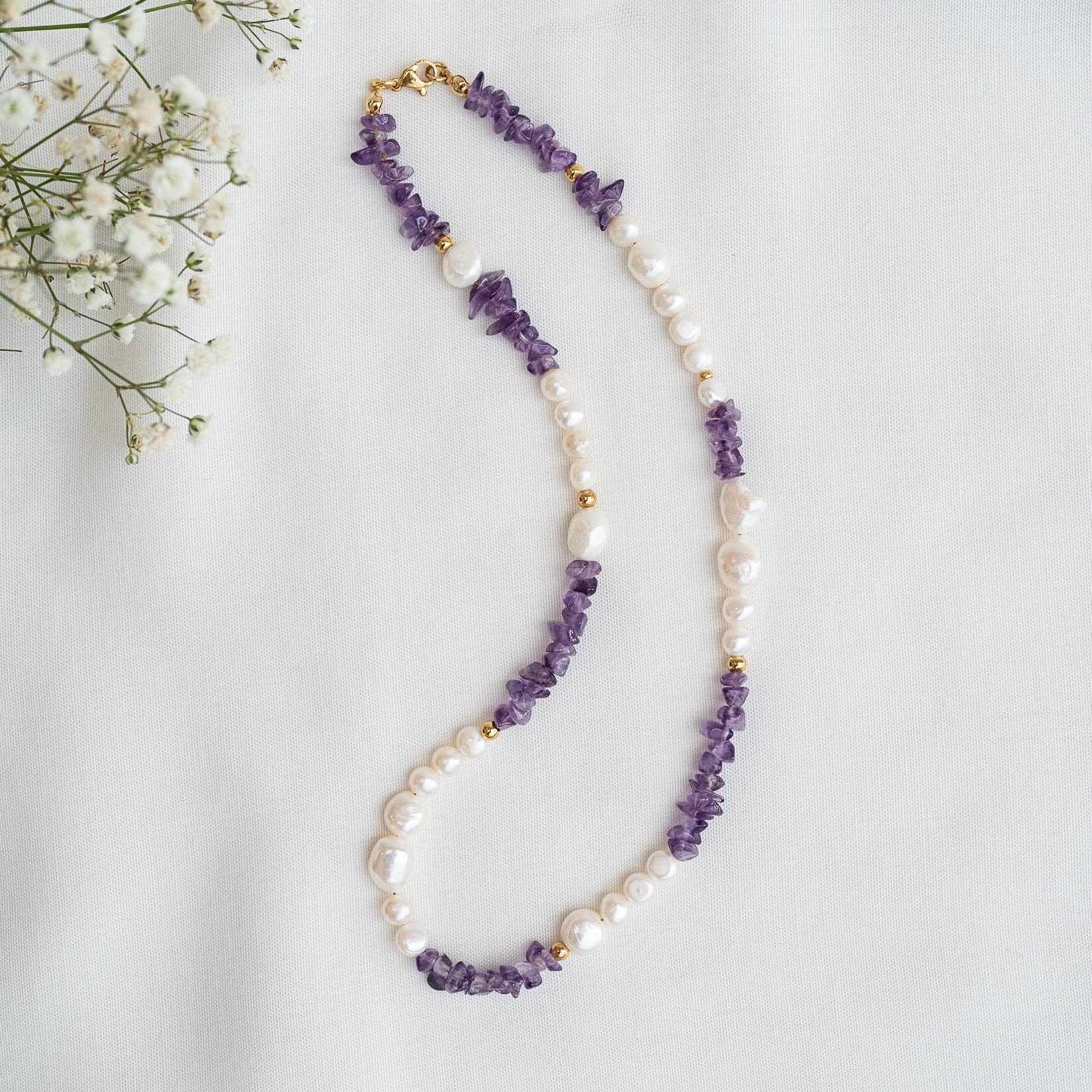 Eine Perlenkette mit abwechselnden Abschnitten aus unregelmäßigen violetten Steinen, weißen Perlen und kleinen Goldperlen ist auf einem weißen Stoffhintergrund abgebildet, mit zarten weißen Blumen in der oberen linken Ecke.