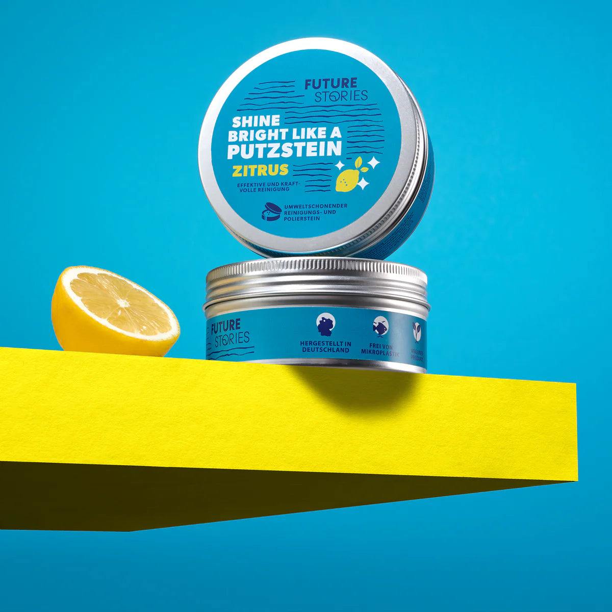 Auf einem gelben Podest vor blauem Hintergrund steht eine Dose Future Stories Zitrus-Reinigungspaste, deren Deckel hochgeklappt ist und neben der eine halbe Zitrone liegt.