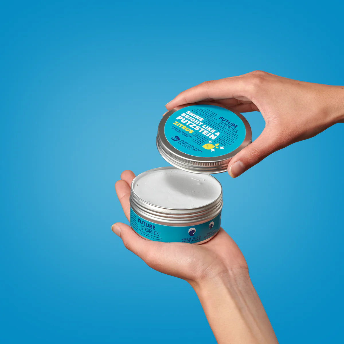 Eine Person öffnet einen runden blauen Behälter mit der Aufschrift Future Stories vor blauem Hintergrund und gibt ein weißes, festes Produkt frei.
