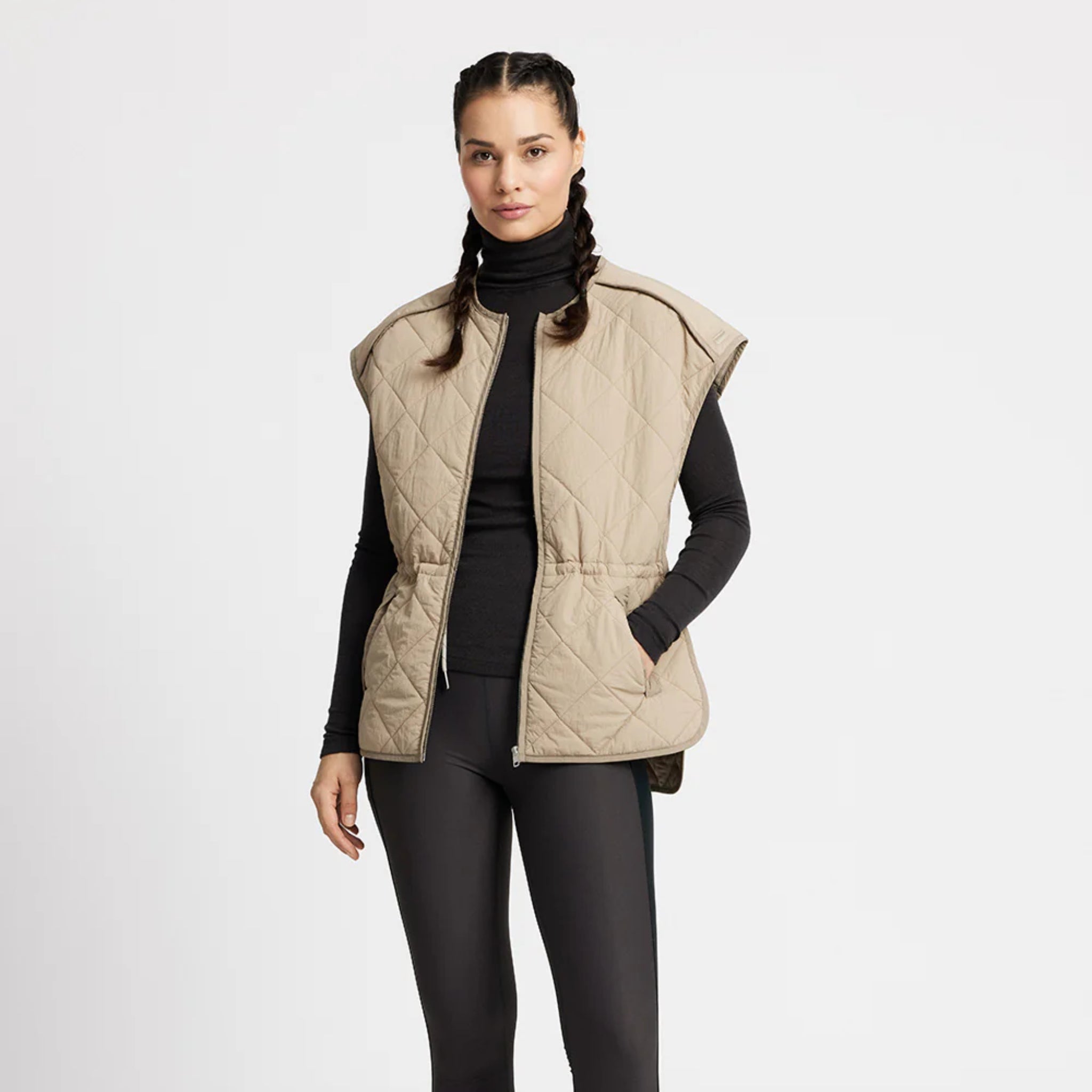 Eine Frau mit geflochtenen Haaren trägt eine beigefarbene Steppweste über einem schwarzen Rollkragenpullover und schwarze Leggings und steht vor einem schlichten weißen Hintergrund.