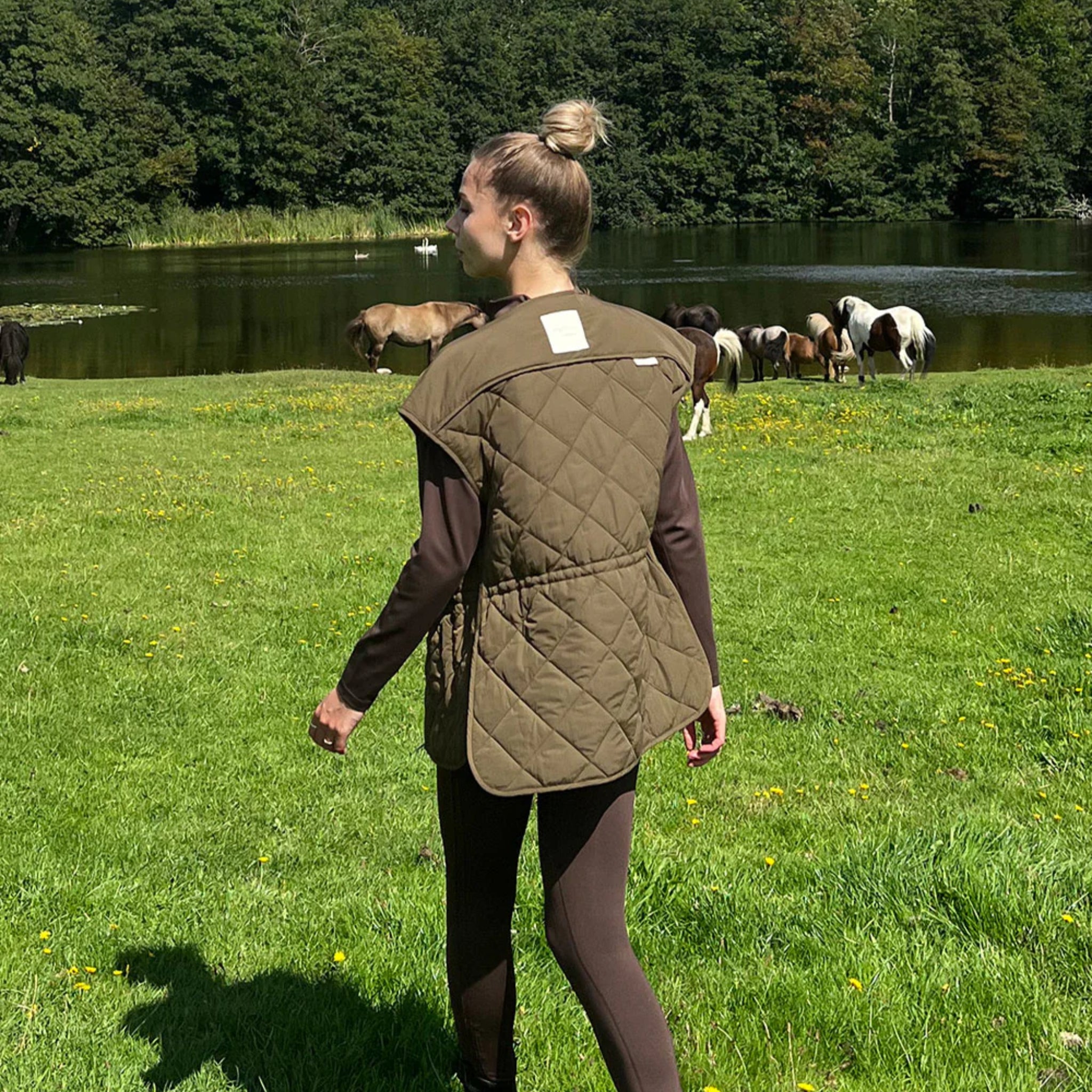 Eine Frau in braunen Leggings und einer Steppweste geht auf grünem Gras in der Nähe eines Teiches spazieren, während im Hintergrund mehrere Pferde grasen und Bäume den Wasserrand säumen.