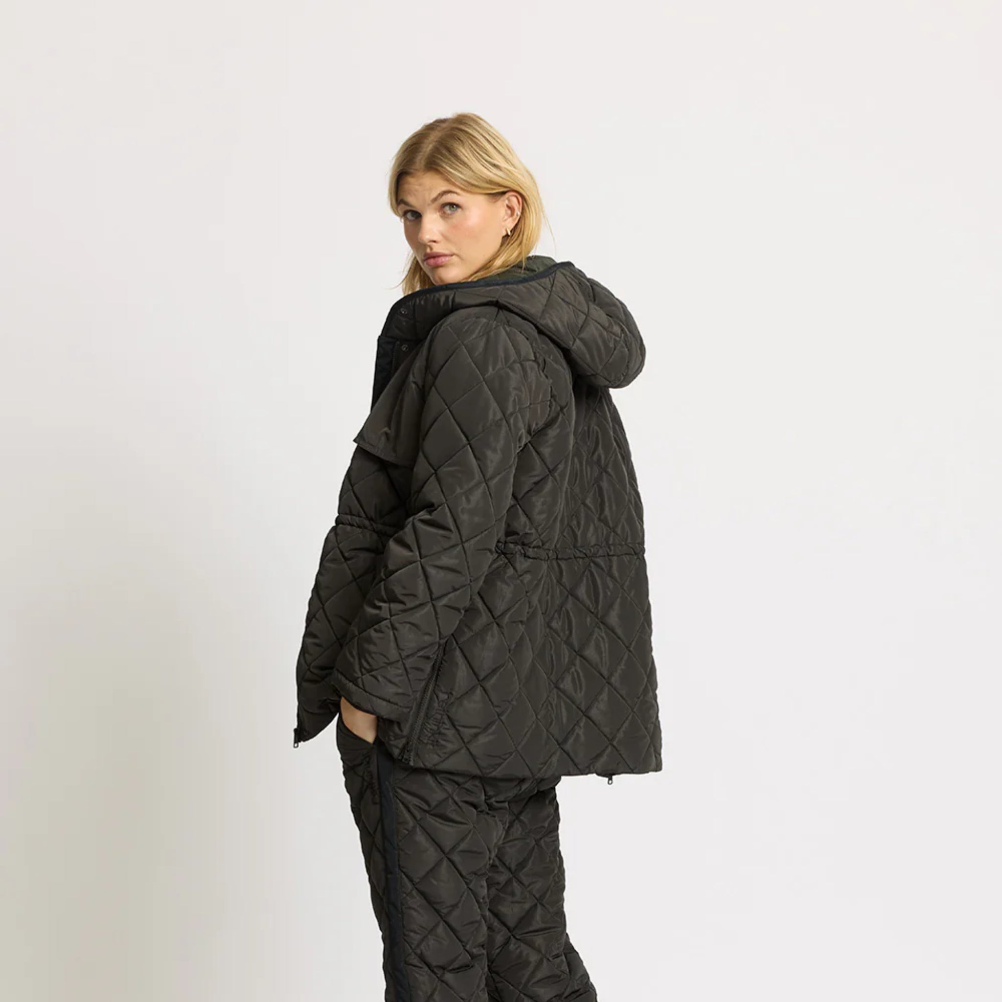 Eine Frau mit blondem Haar trägt eine dunkle Steppjacke mit Kapuze und eine dazu passende Hose. Sie steht seitlich und schaut über ihre Schulter vor einem schlichten hellen Hintergrund.