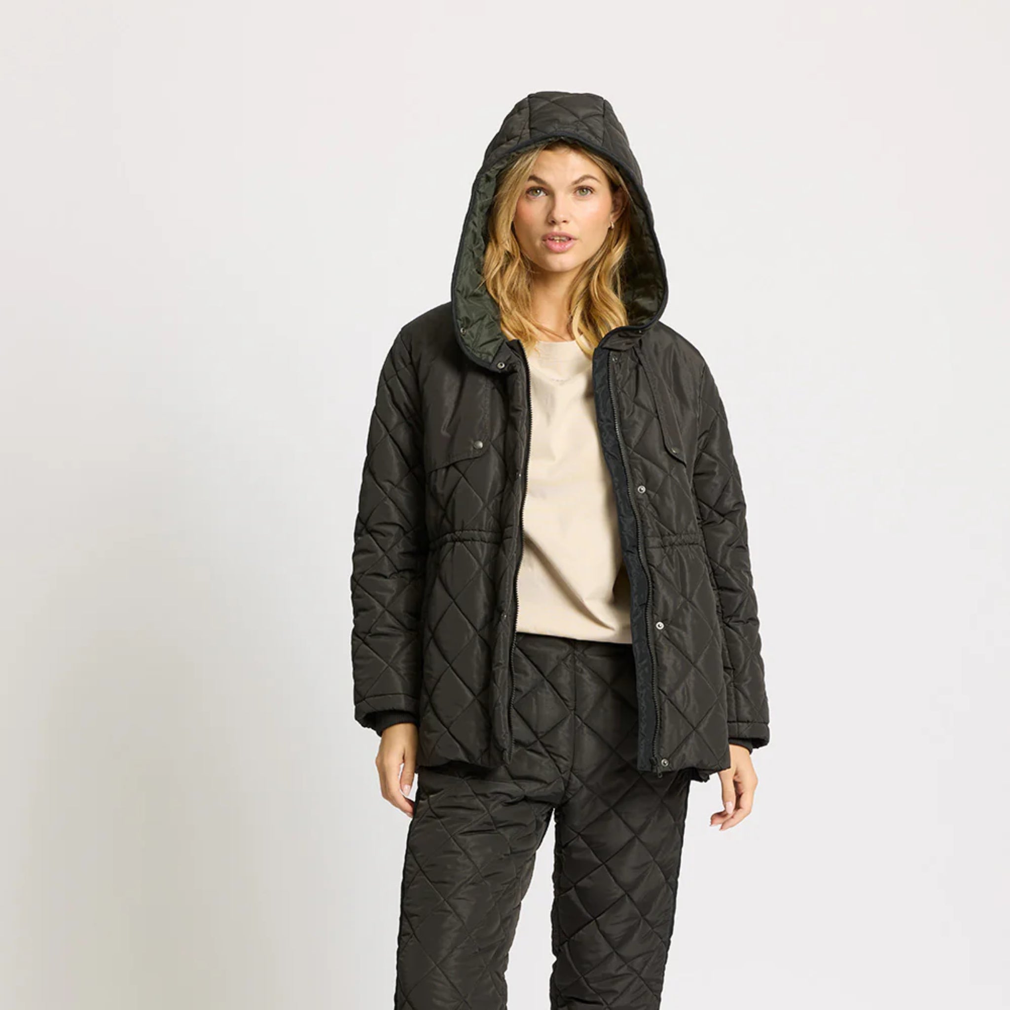 Eine Frau, die eine gesteppte schwarze Kapuzenjacke und eine dazu passende gesteppte Hose trägt, steht vor einem schlichten hellen Hintergrund und blickt mit neutralem Blick nach vorn.