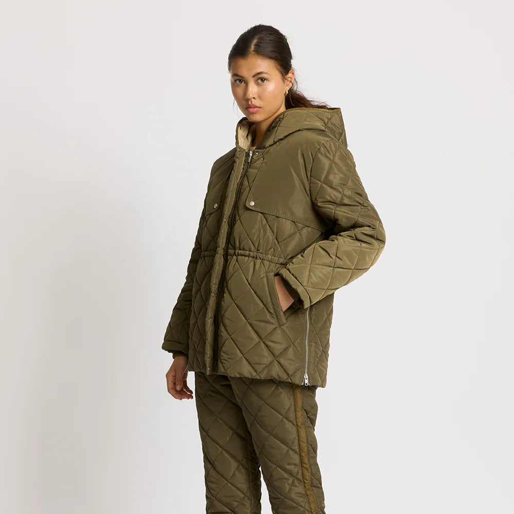 Eine Frau steht vor einem schlichten, hellen Hintergrund und trägt eine olivgrüne Steppjacke mit Kapuze und eine dazu passende Stepphose. Sie schaut leicht zur Seite und hat die Hände in den Jackentaschen.
