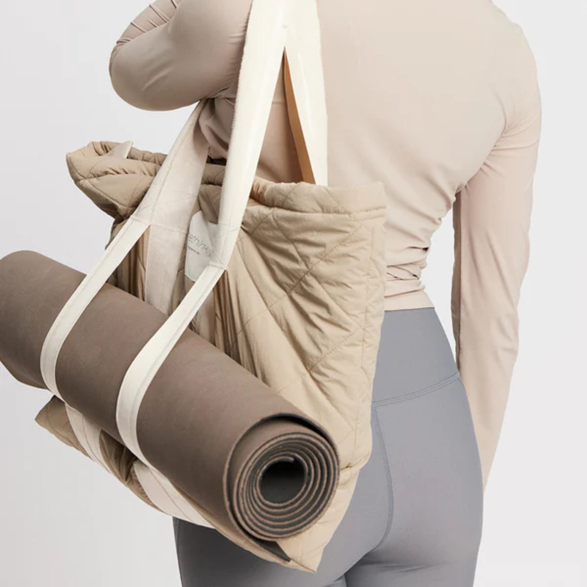 Person trägt eine beigefarbene Tasche mit weißem Henkel, an der eine aufgerollte Yogamatte befestigt ist.