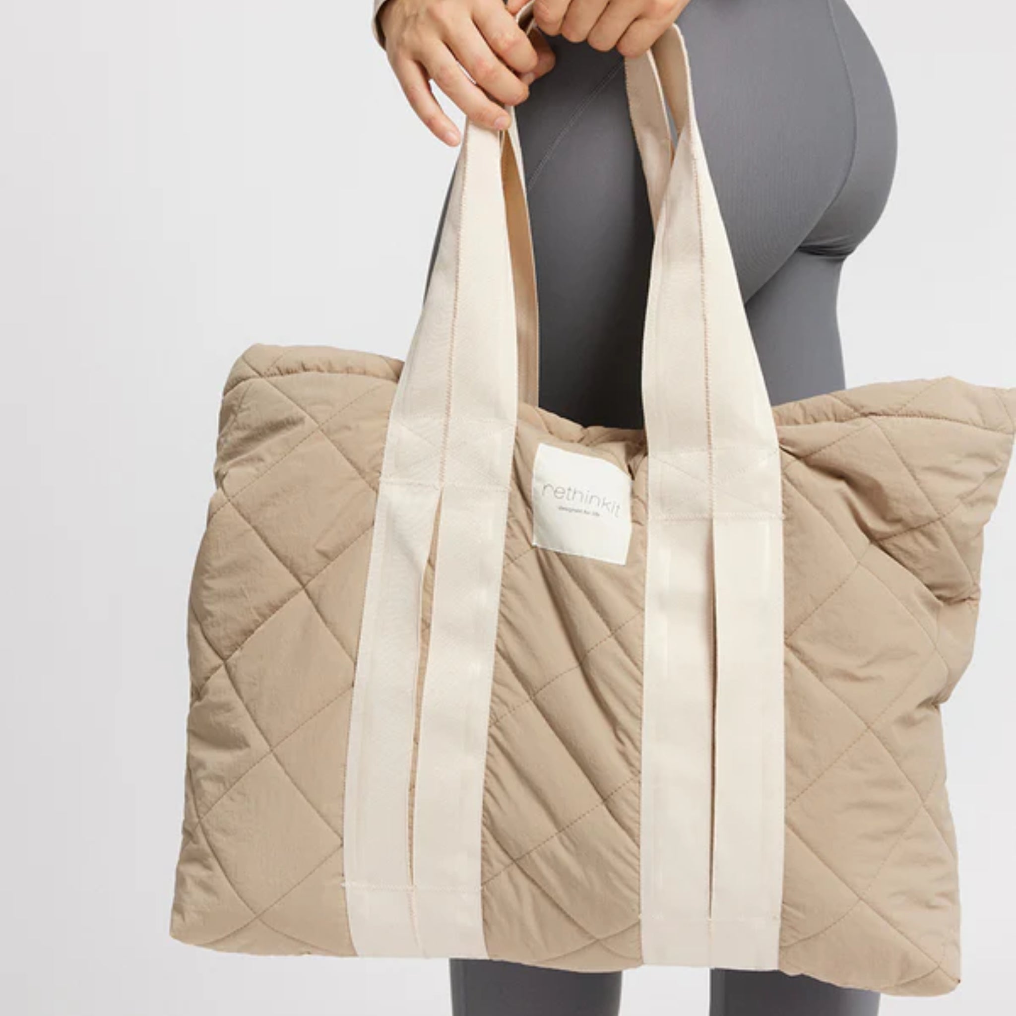 Person trägt eine gesteppte, beige Tragetasche aus Stoff mit weißen Henkeln und einem Label.