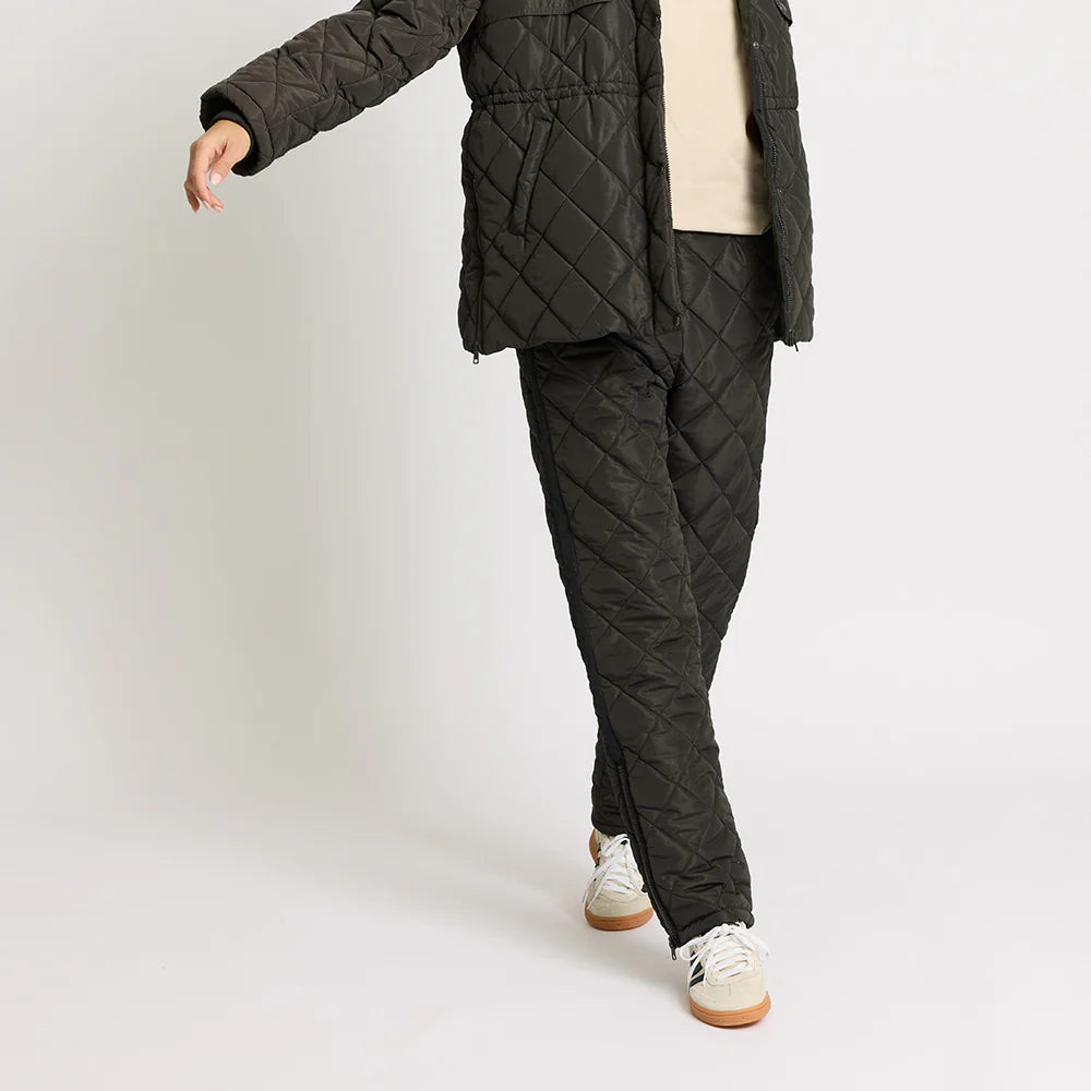 Eine Frau posiert in einer gesteppten schwarzen Jacke und einer passenden gesteppten Hose, mit einem beigen Hemd und weißen Turnschuhen, vor einem schlichten weißen Hintergrund.