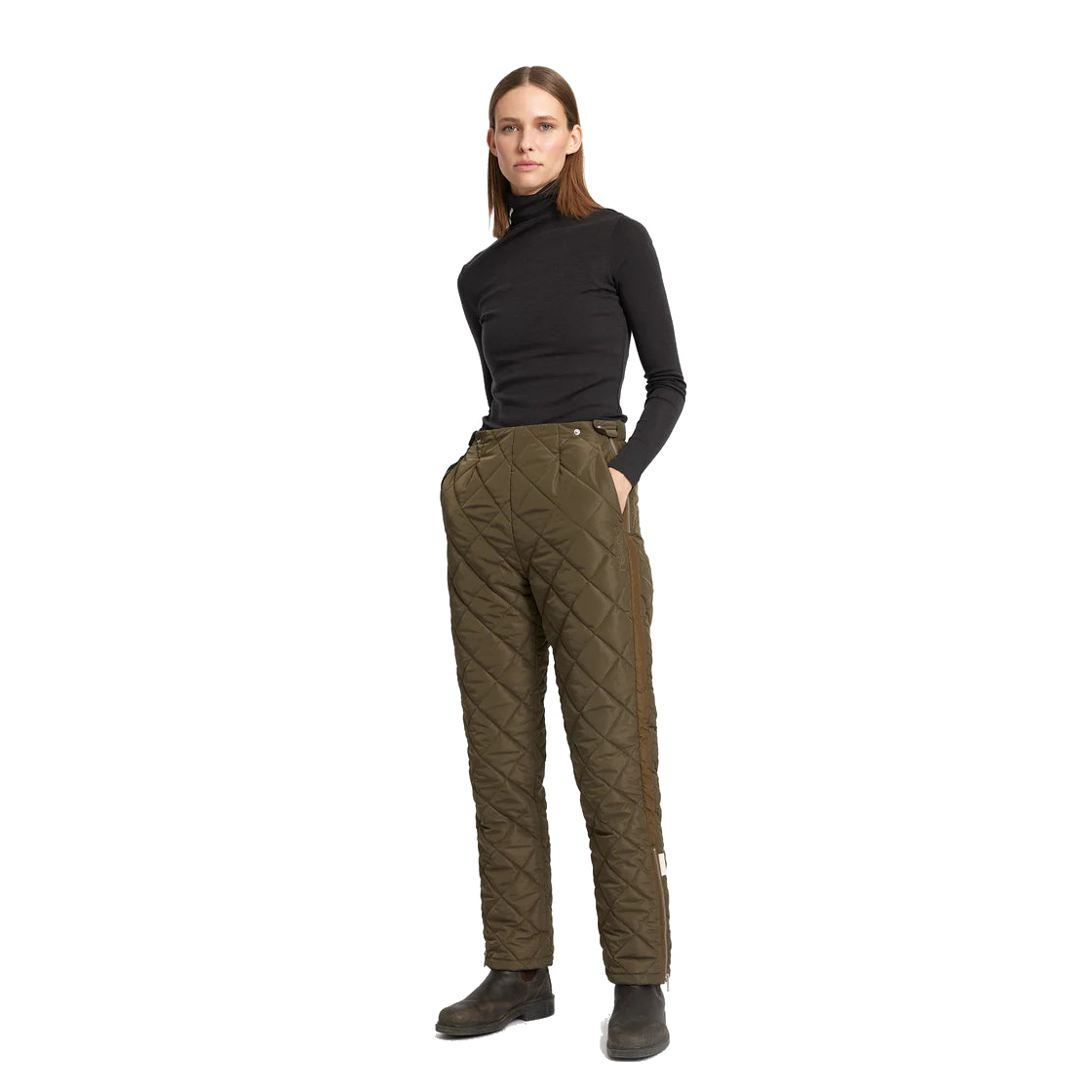 Eine Frau steht vor einem weißen Hintergrund, trägt einen schwarzen Rollkragenpullover, eine olivgrüne Stepphose und schwarze Stiefel und hat die Hände in den Taschen.