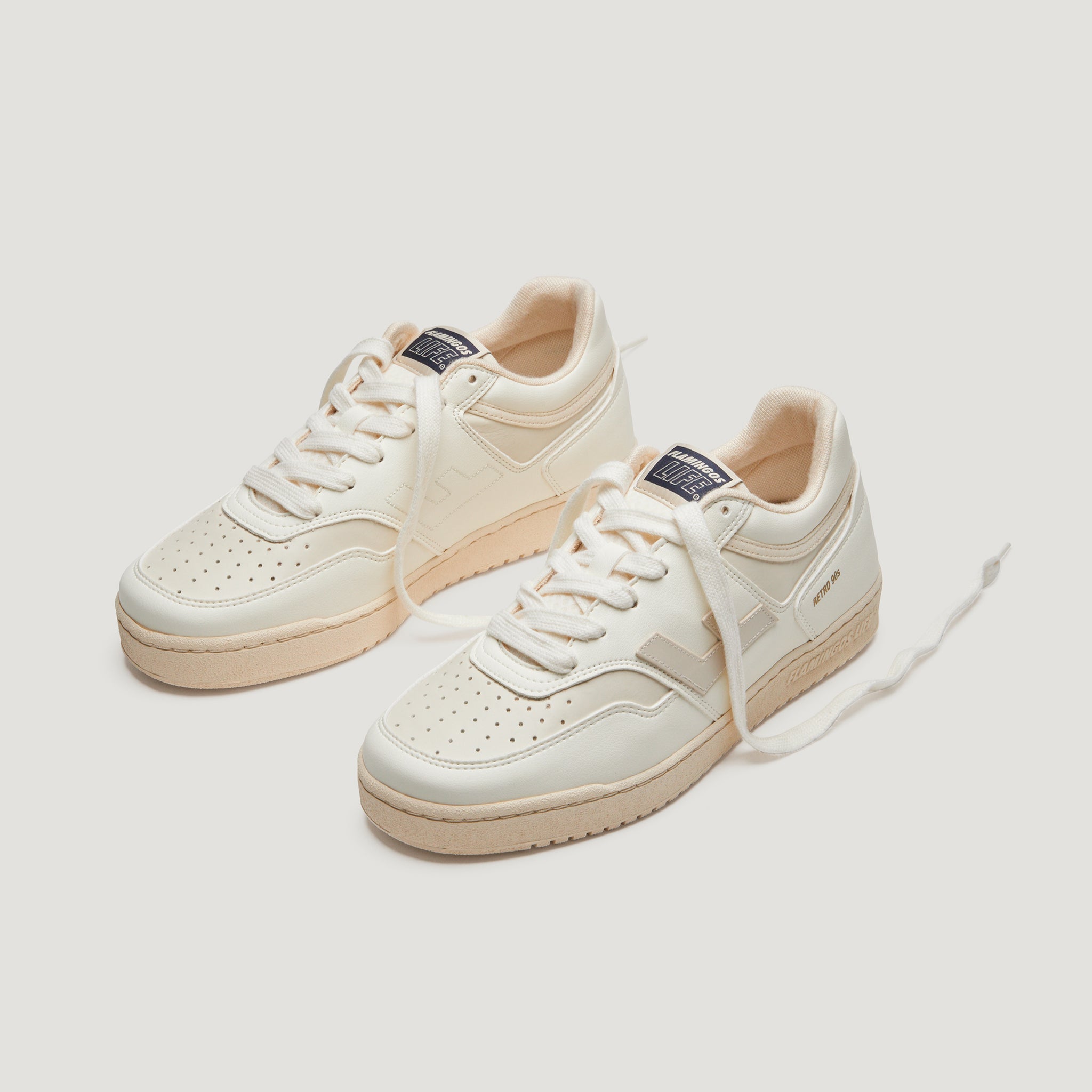 Weiße Sneakers mit beiger Sohle und dunkelblauen Details, auf hellem Hintergrund, die Schnürsenkel teilweise offen.