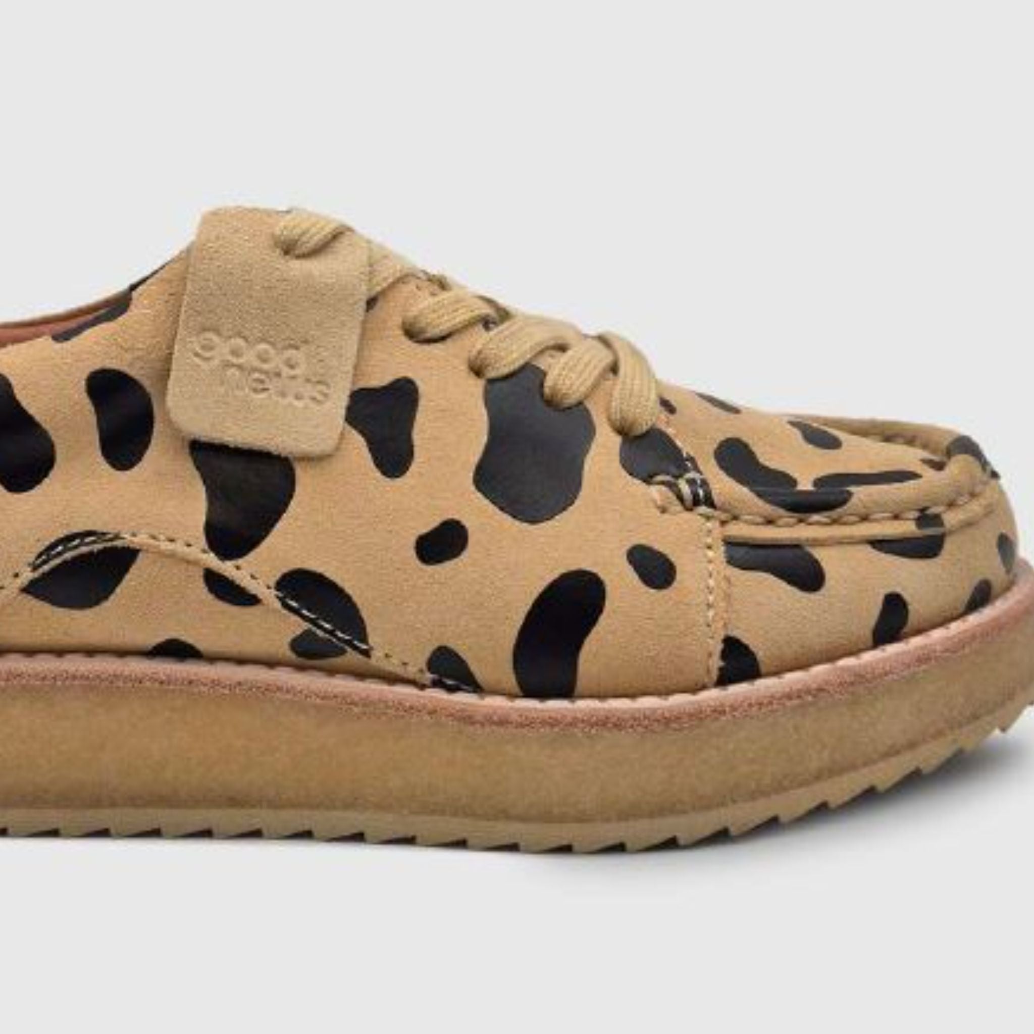 Ein hellbrauner Wildlederschuh mit schwarzen, unregelmäßigen leopardenähnlichen Flecken, hellbraunen Schnürsenkeln, einer dicken hellbraunen Sohle und einem kleinen Etikett an der Seite, auf dem gute Nachrichten stehen.