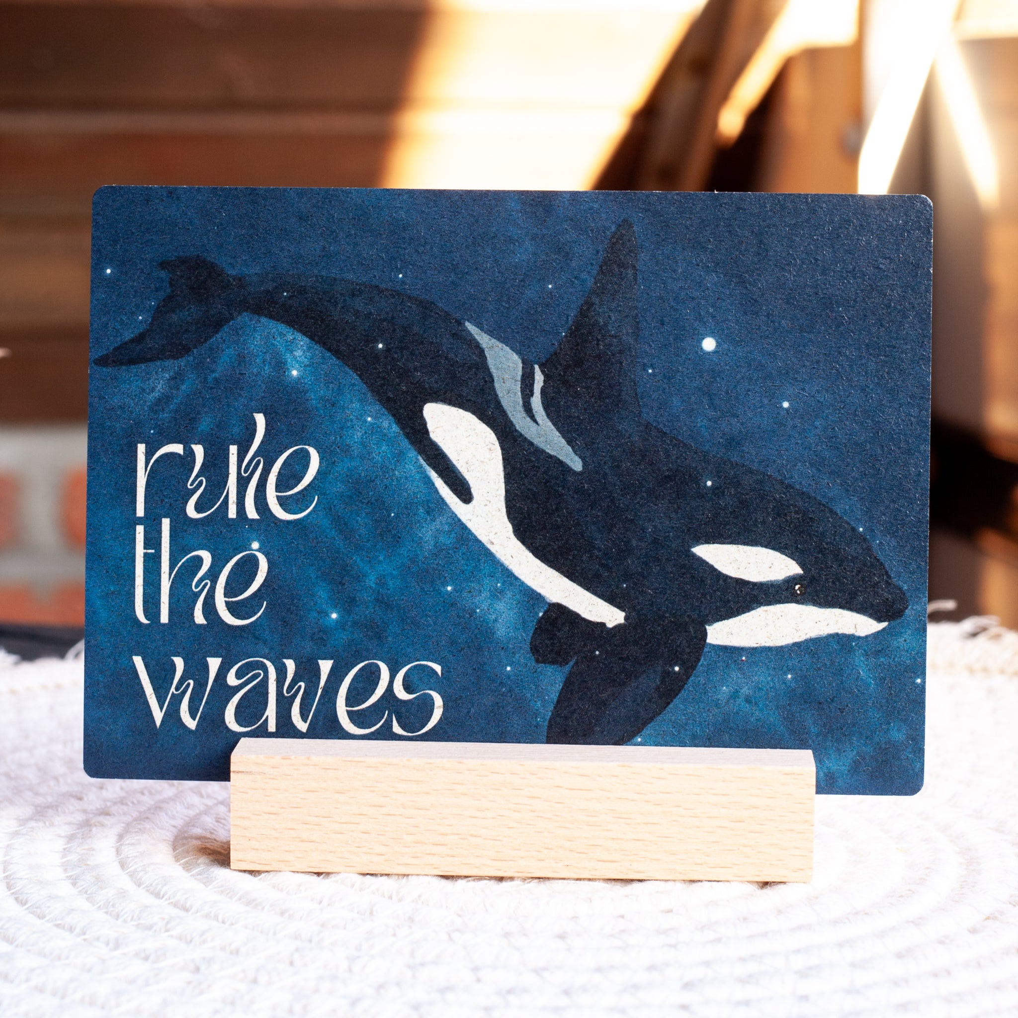 Eine Postkarte mit der Illustration eines Orcas, der in einem sternenklaren blauen Meer schwimmt, auf einem Holzständer stehend. Der Text auf der Karte lautet "rule the waves" in weißen Buchstaben.