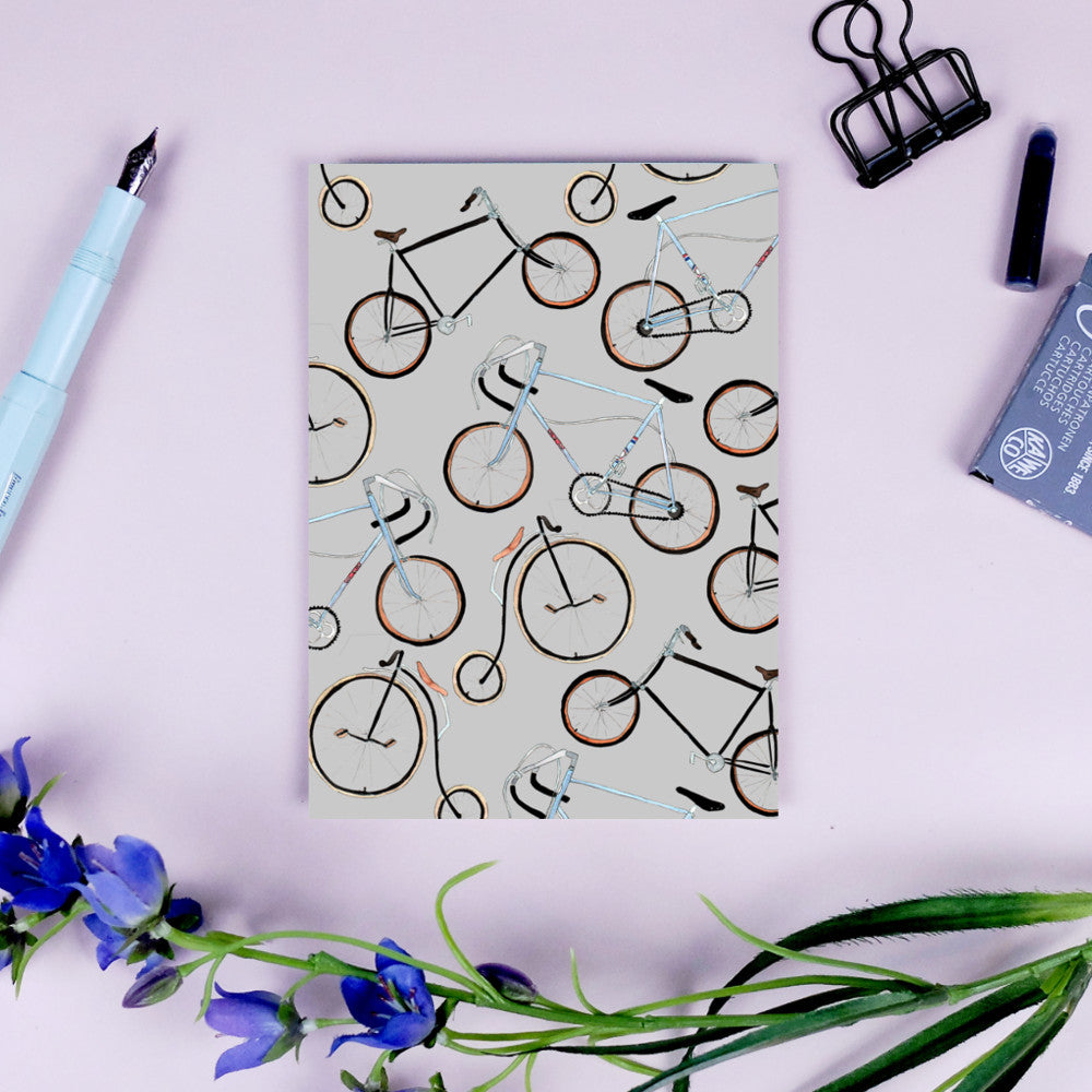 Papier mit Fahrradmuster, umgeben von einem Stift, Büroklammer und Blumen auf einem rosa Hintergrund.