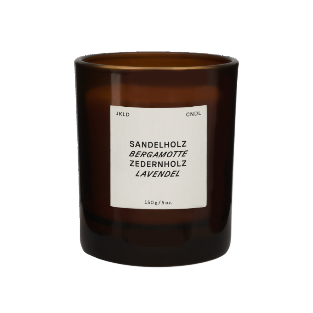 Braune Duftkerze im Glas mit Etikett, das Sandelholz, Bergamotte, Zedernholz und Lavendel als Duftstoffe auflistet.