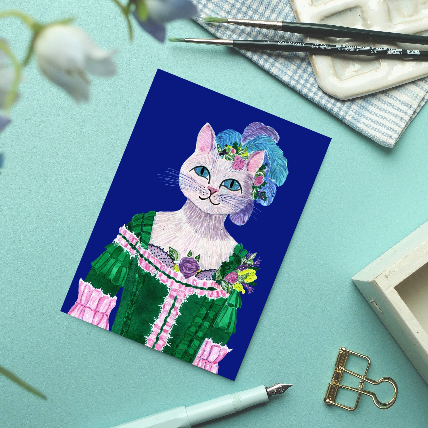Eine Postkarte mit der Illustration einer weißen Katze in einem verzierten grün-rosa Kleid im viktorianischen Stil, mit einem Blumenkopfschmuck, die auf einer blauen Fläche neben Kunstzubehör und einer goldenen Büroklammer liegt.