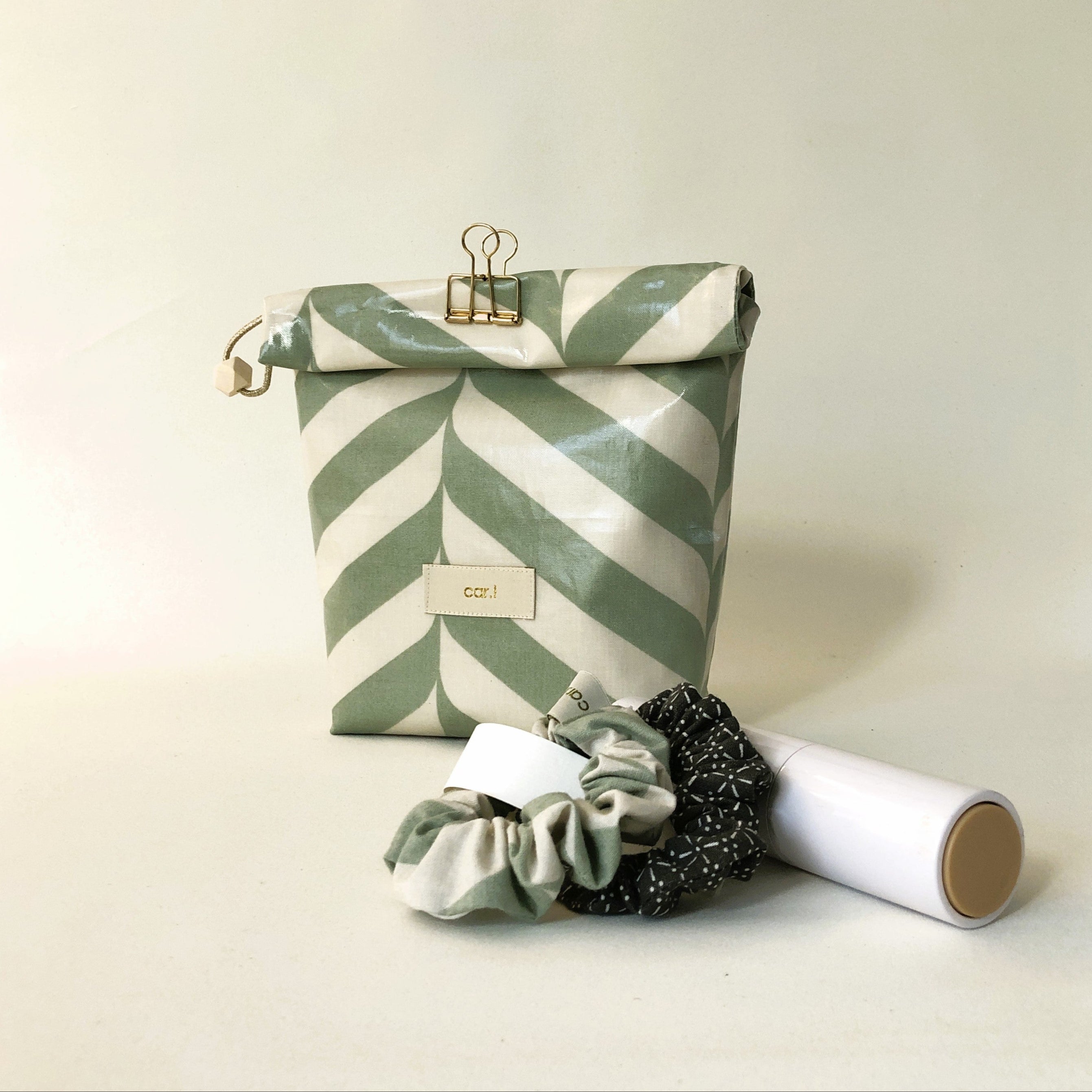 Mehrfarbige Tasche mit Blattmuster, zwei Haargummis und eine weiße zylindrische Verpackung auf hellem Hintergrund.