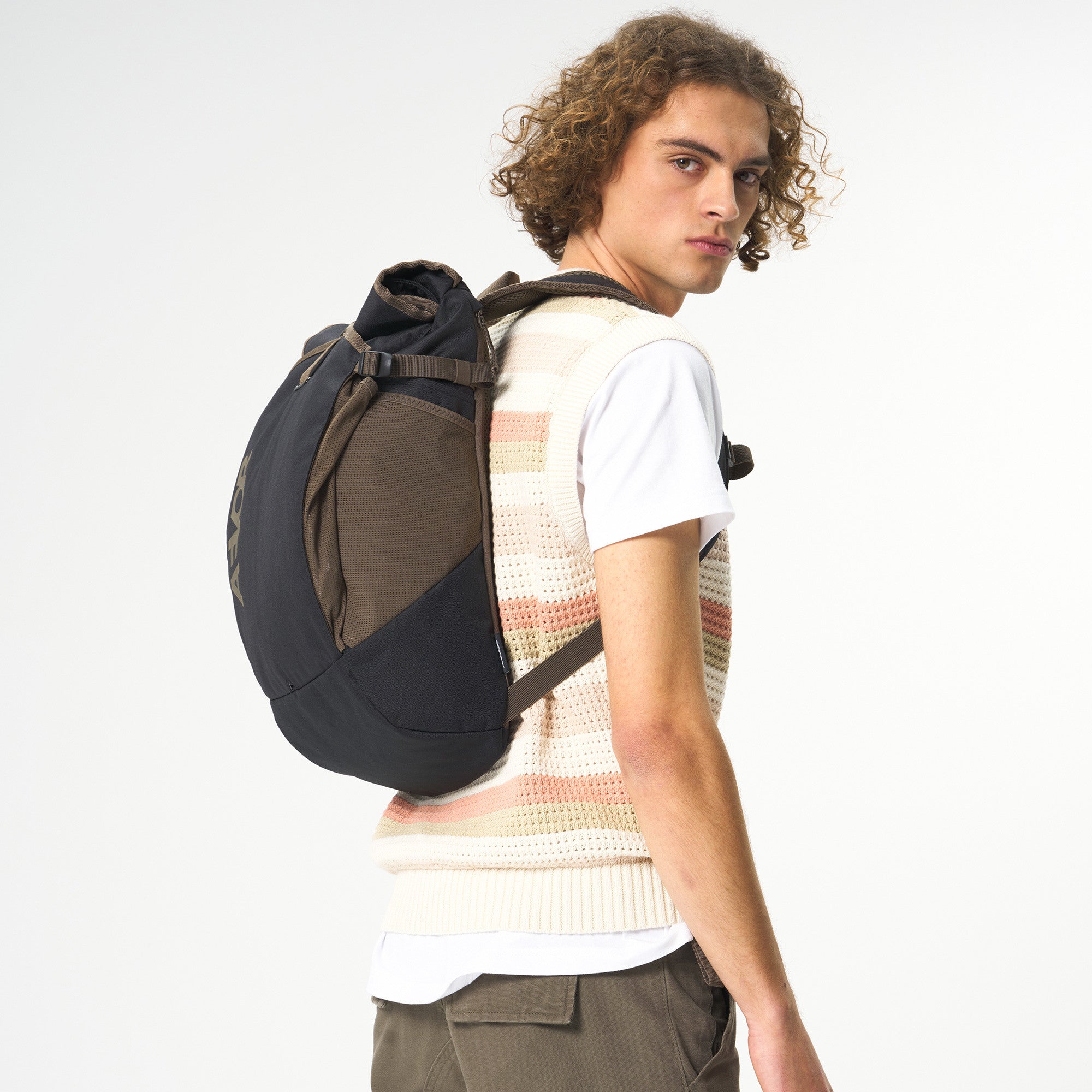 Ein junger Mann mit lockigem Haar, der eine ärmellose Strickweste über einem weißen T-Shirt und eine olivfarbene Hose trägt, blickt über die Schulter, während er einen dunkelfarbigen Rucksack trägt. Der Hintergrund ist einfarbig weiß.