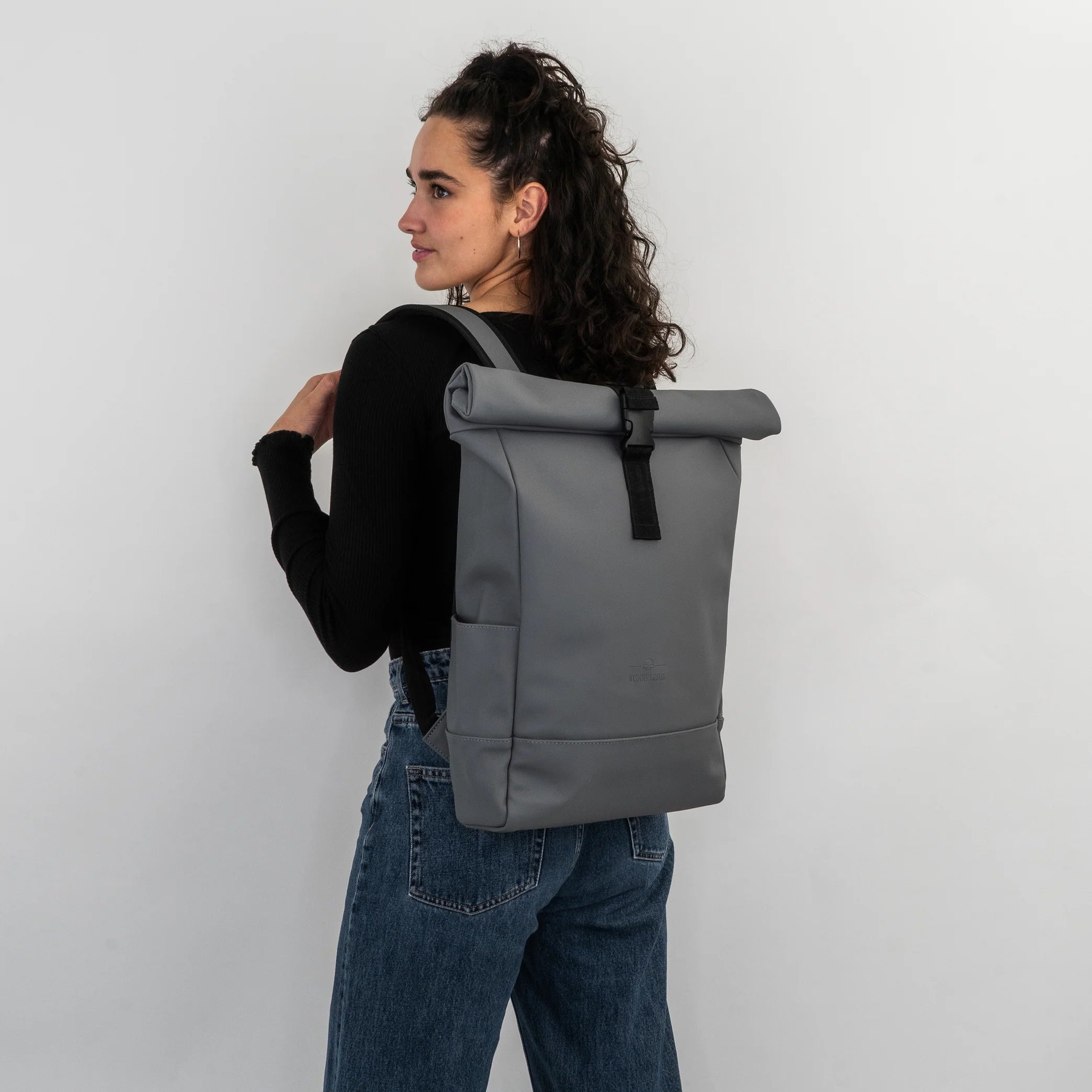 Eine Frau mit lockigem Haar, die ein schwarzes, langärmeliges Oberteil und blaue Jeans trägt, steht seitlich und schaut über ihre Schulter, während sie einen großen, grauen Rolltop-Rucksack vor einem schlichten weißen Hintergrund modelliert.