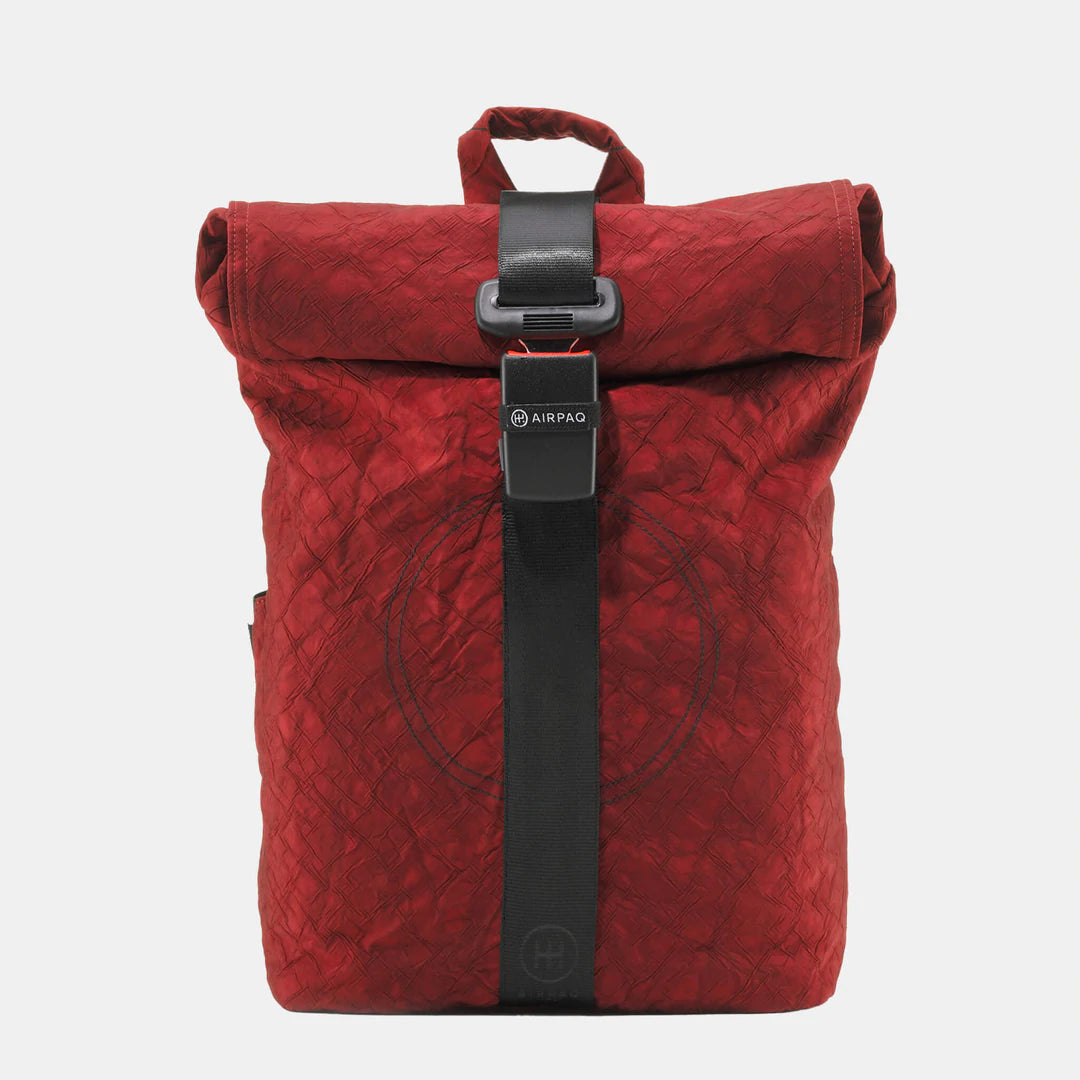 Ein roter Rolltop-Rucksack mit schwarzem, vertikalem Riemen und Schnallenverschluss, der auf der Vorderseite ein dezentes, kreisförmiges Muster und auf der Schnalle ein AIRPAQ-Label aufweist. Der Hintergrund ist einfarbig weiß.