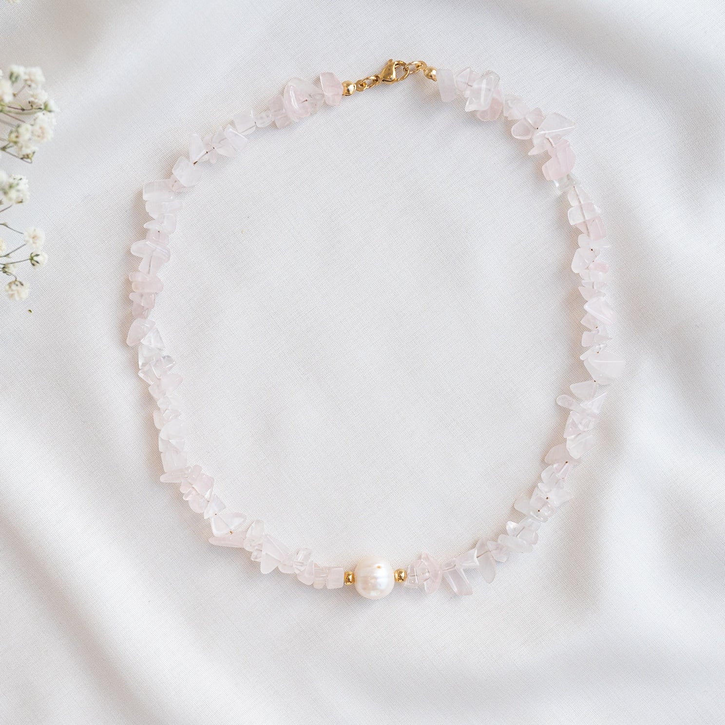 Eine Halskette aus blassrosa Edelsteinchips mit einer einzelnen großen weißen Perle in der Mitte, die auf einem weißen Stoffgrund ruht. Oben ist ein kleiner Goldverschluss zu sehen.