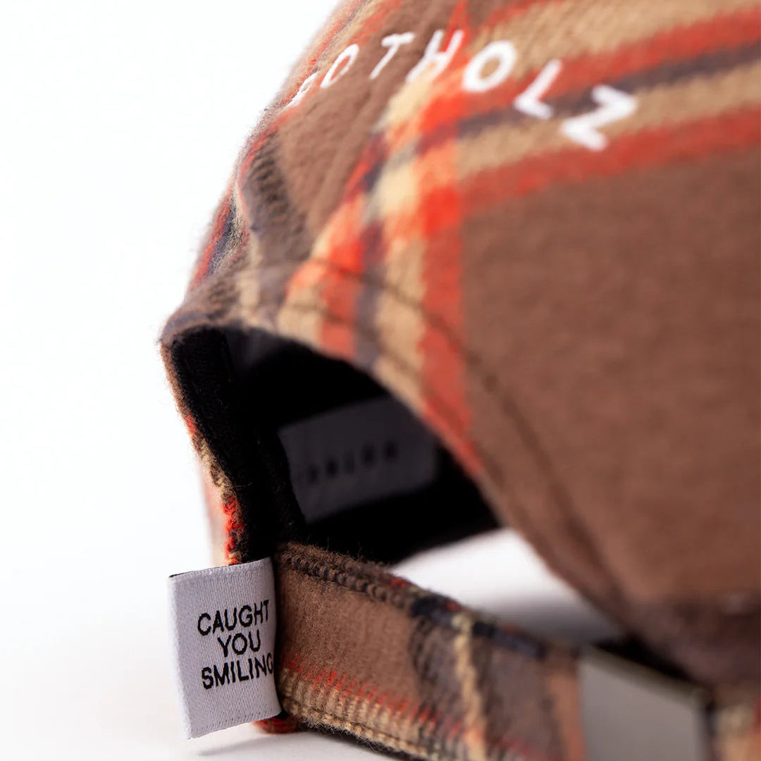 Nahaufnahme einer braun und orange karierten Mütze mit weißer Stickerei. Ein kleines Stoffschild in der Nähe des verstellbaren Riemens trägt die Aufschrift "CAUGHT YOU SMILING" in schwarzer Schrift.