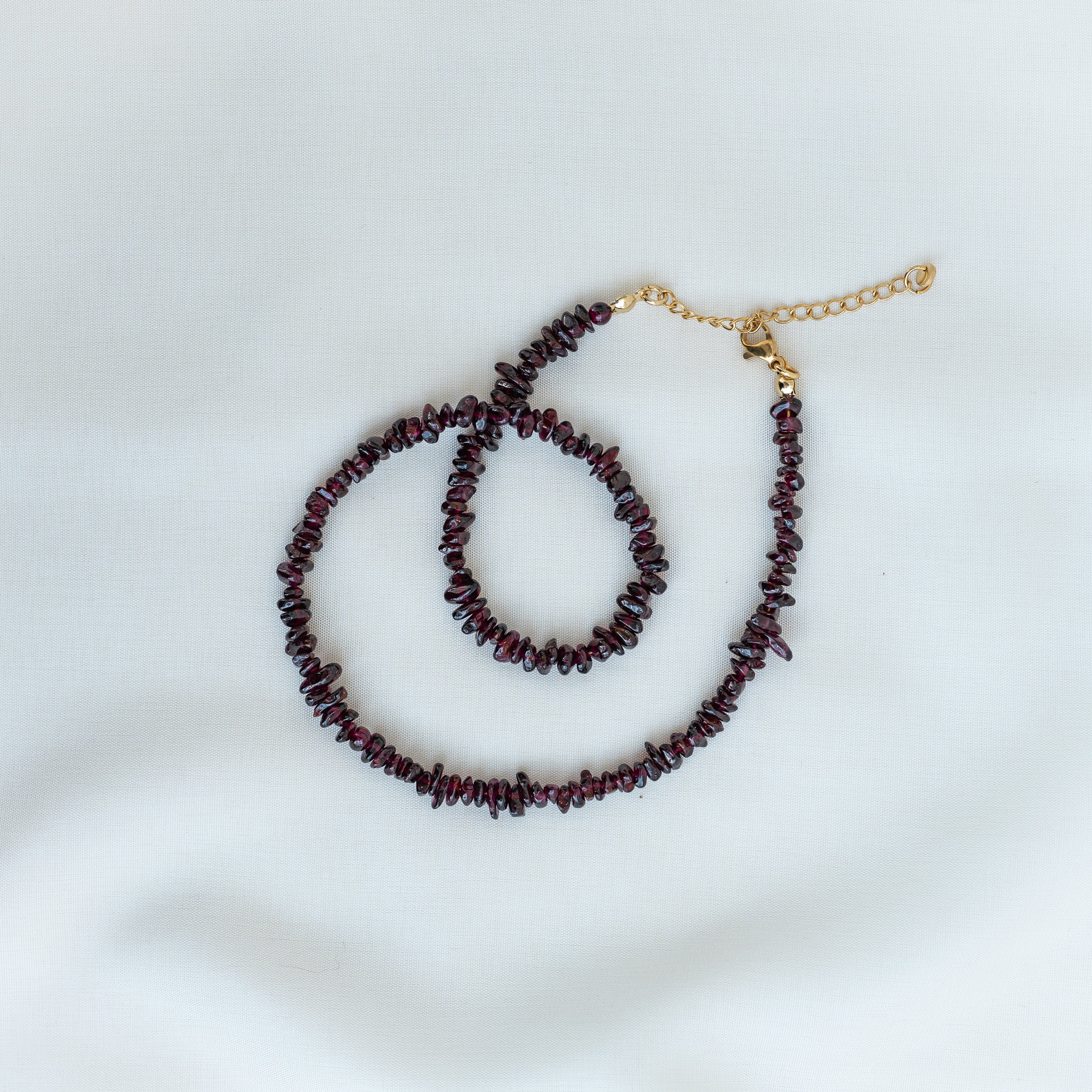 Eine Halskette mit dunkelroten, unregelmäßigen Edelsteinperlen und einem zarten goldenen Verschluss und Verlängerungsstück ist auf einem hellen, neutralen Hintergrund aufgezogen.