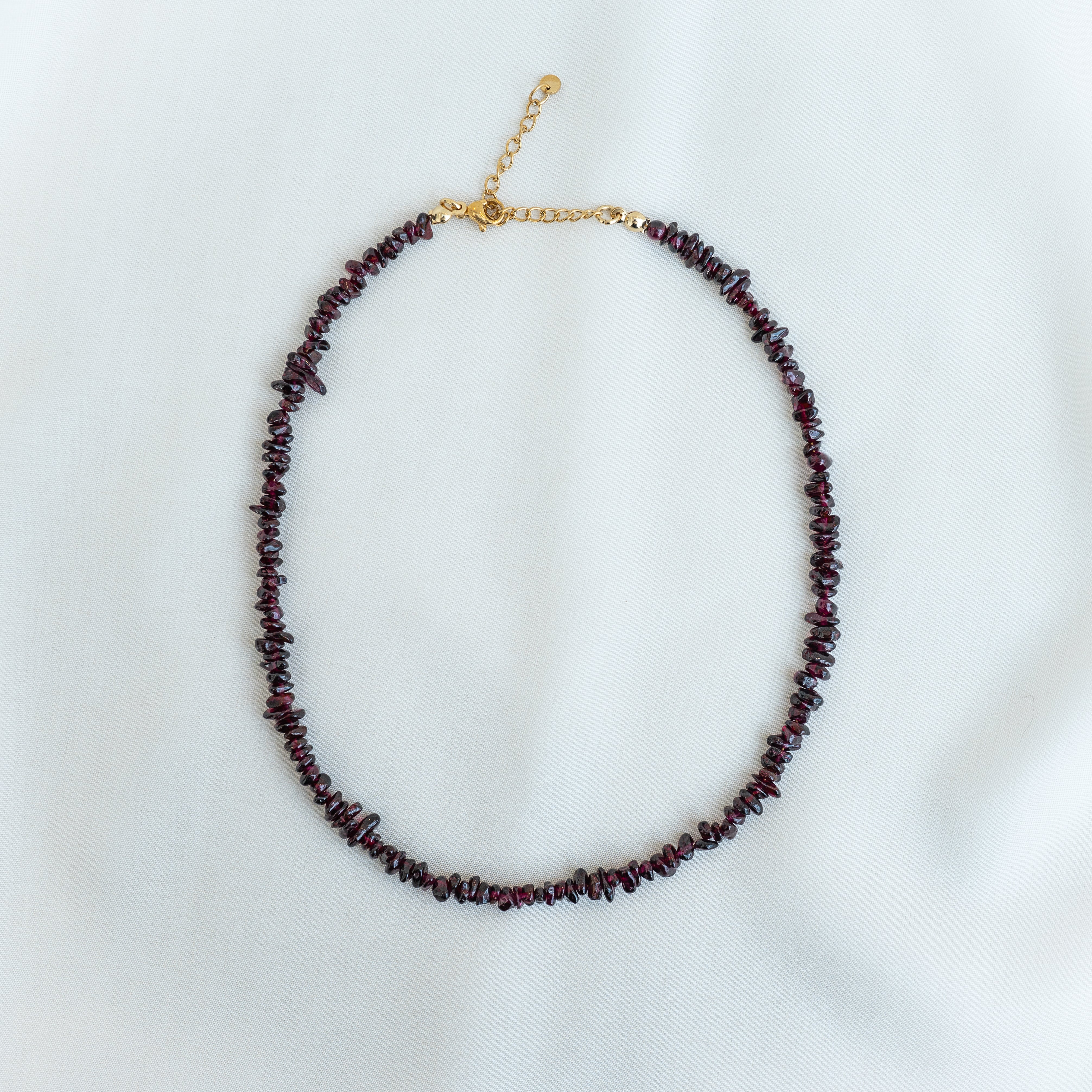 Eine Perlenkette mit unregelmäßigen dunkelroten Steinen und einem goldenen Verschluss und einer Verlängerungskette, die auf einem weißen Stoffhintergrund angebracht ist.