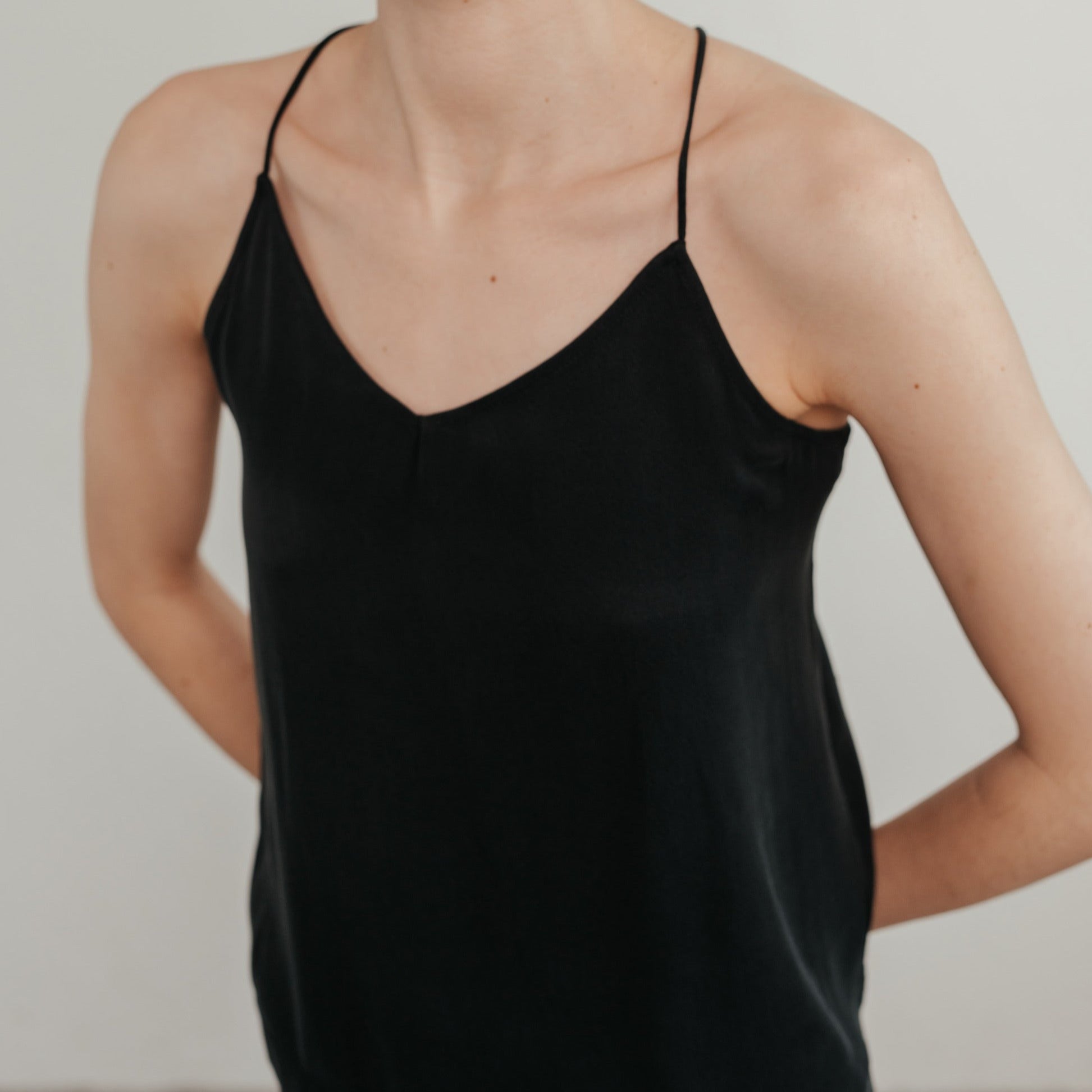 Person in einem schwarzen Spaghettiträger-Top vor neutralem Hintergrund, mit nach hinten gehaltenen Armen.