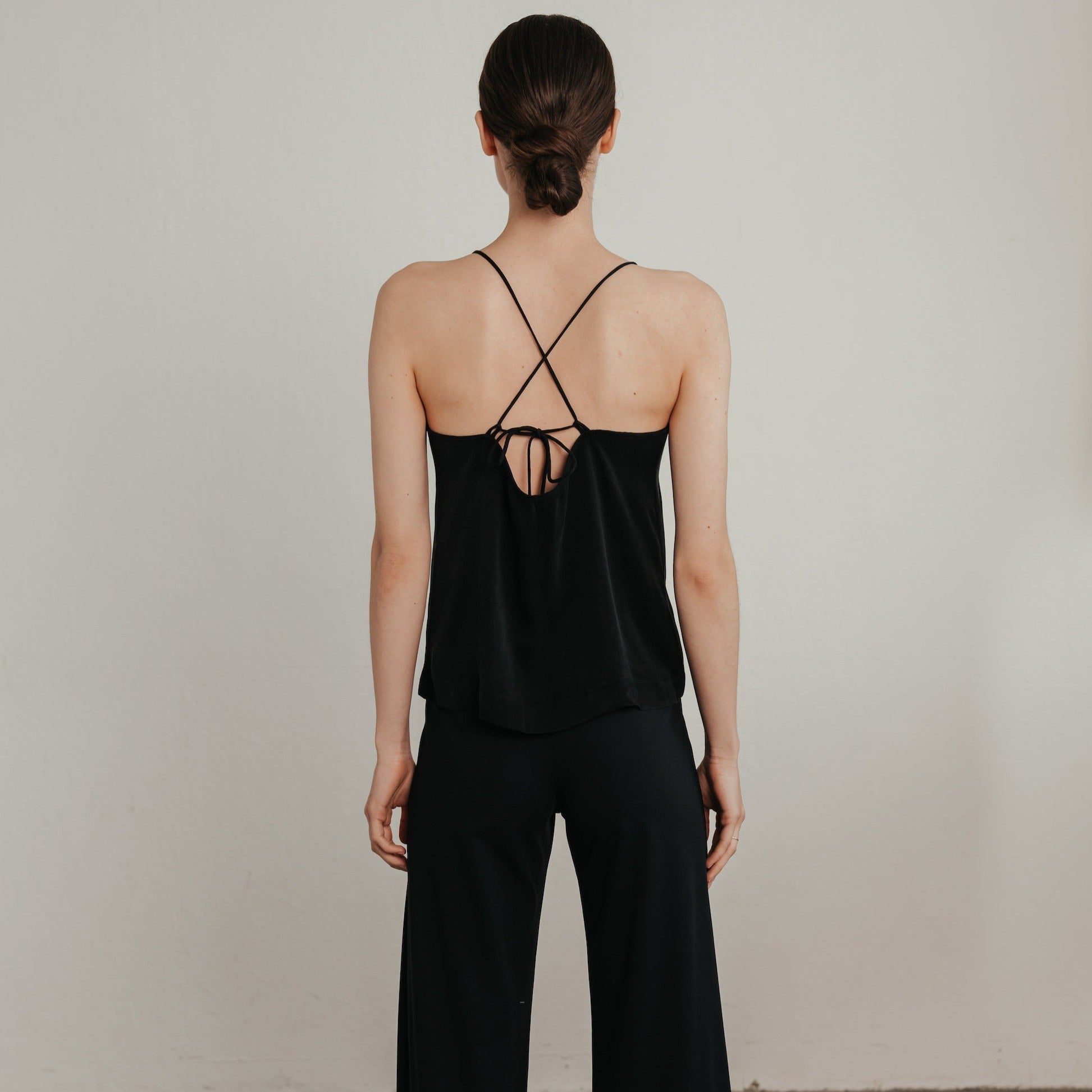 Frau mit zusammengebundenem Haar trägt ein schwarzes Oberteil mit Kreuzträgern und schwarze Hosen, von hinten gesehen.