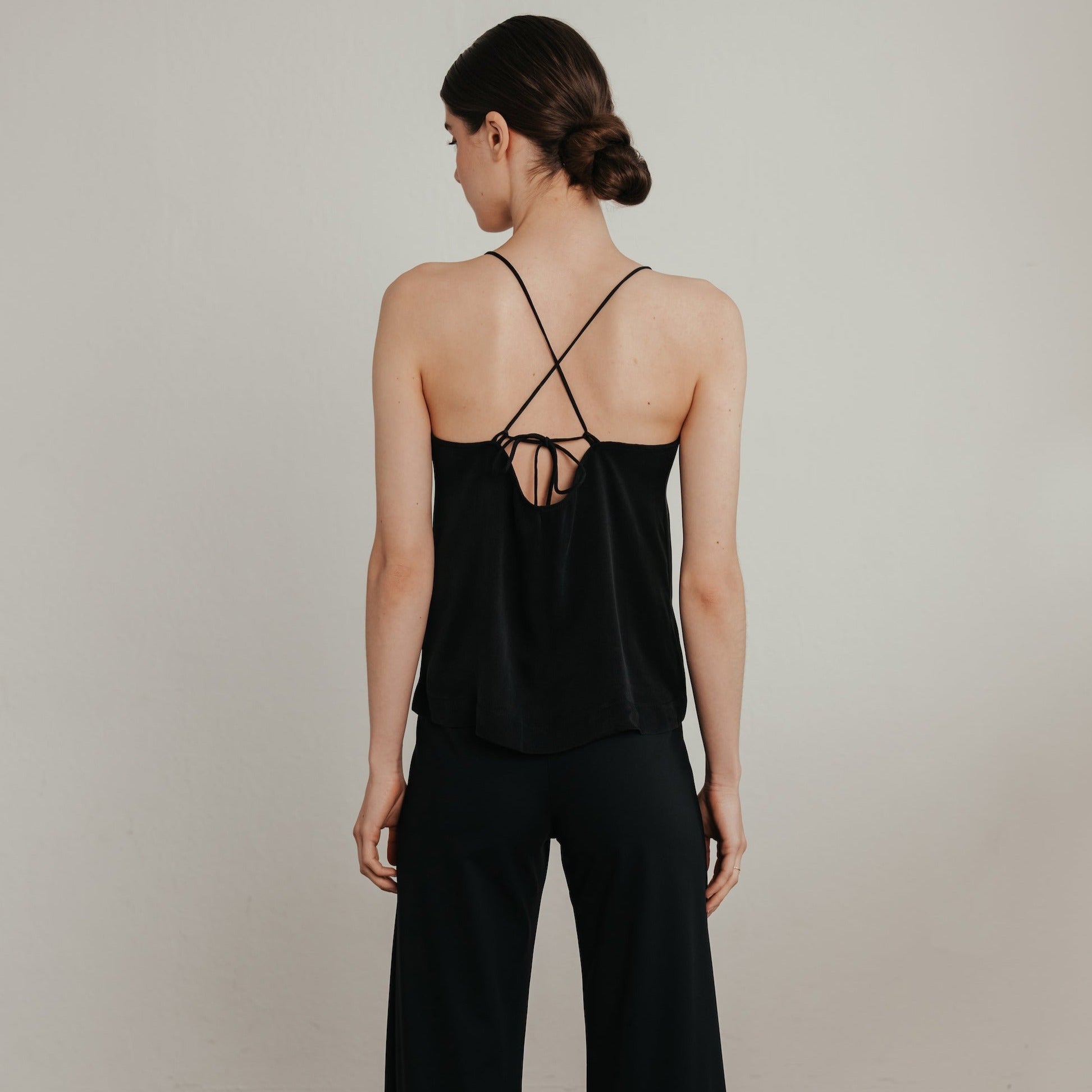 Frau mit zurückgestecktem Haar trägt ein schwarzes Oberteil mit dünnen Trägern, die sich am Rücken überkreuzen.