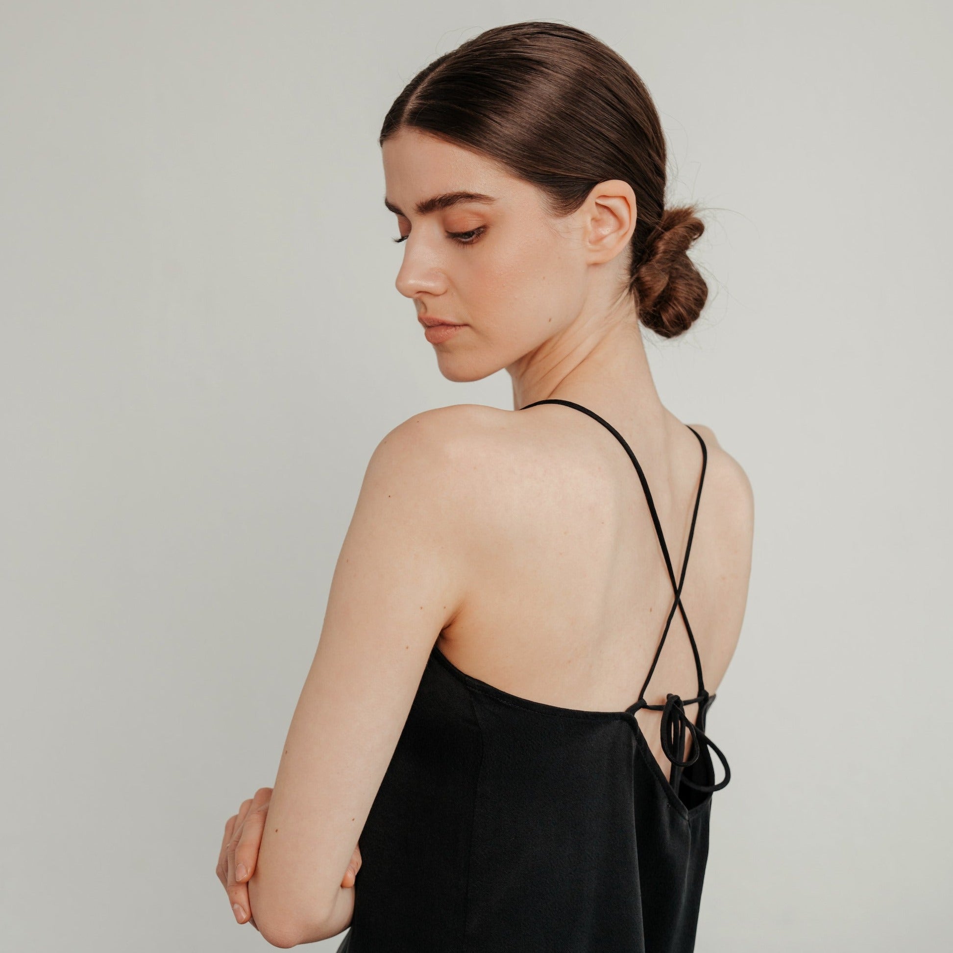 Frau in schwarzem Oberteil mit gekreuzten Trägern, blickt zur Seite, Haare zum Knoten gebunden.