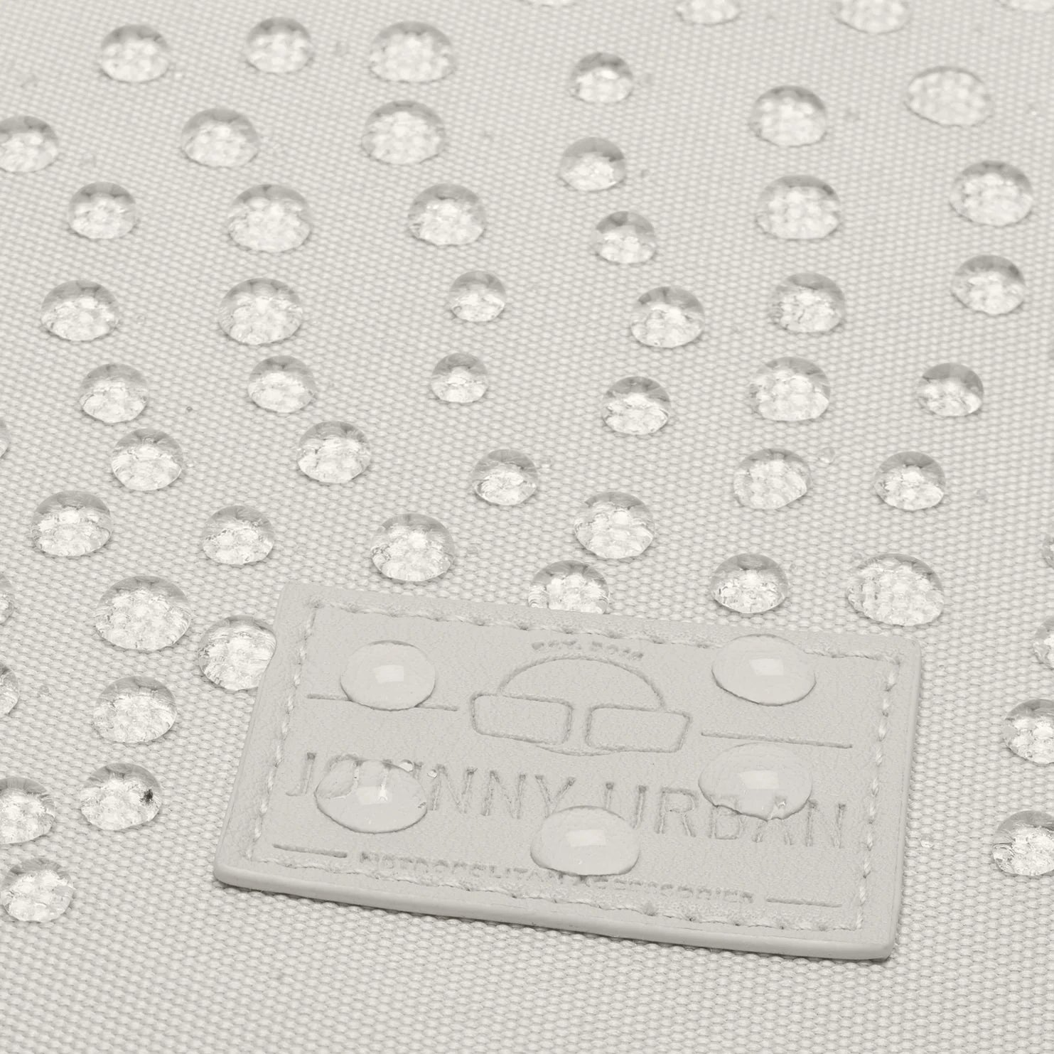 Nahaufnahme einer hellen Stoffoberfläche mit Wassertropfen und einem geprägten Etikett mit der Aufschrift "Johnny Urban". Das Etikett ist leicht feucht, und die Textur des Stoffes ist sichtbar.